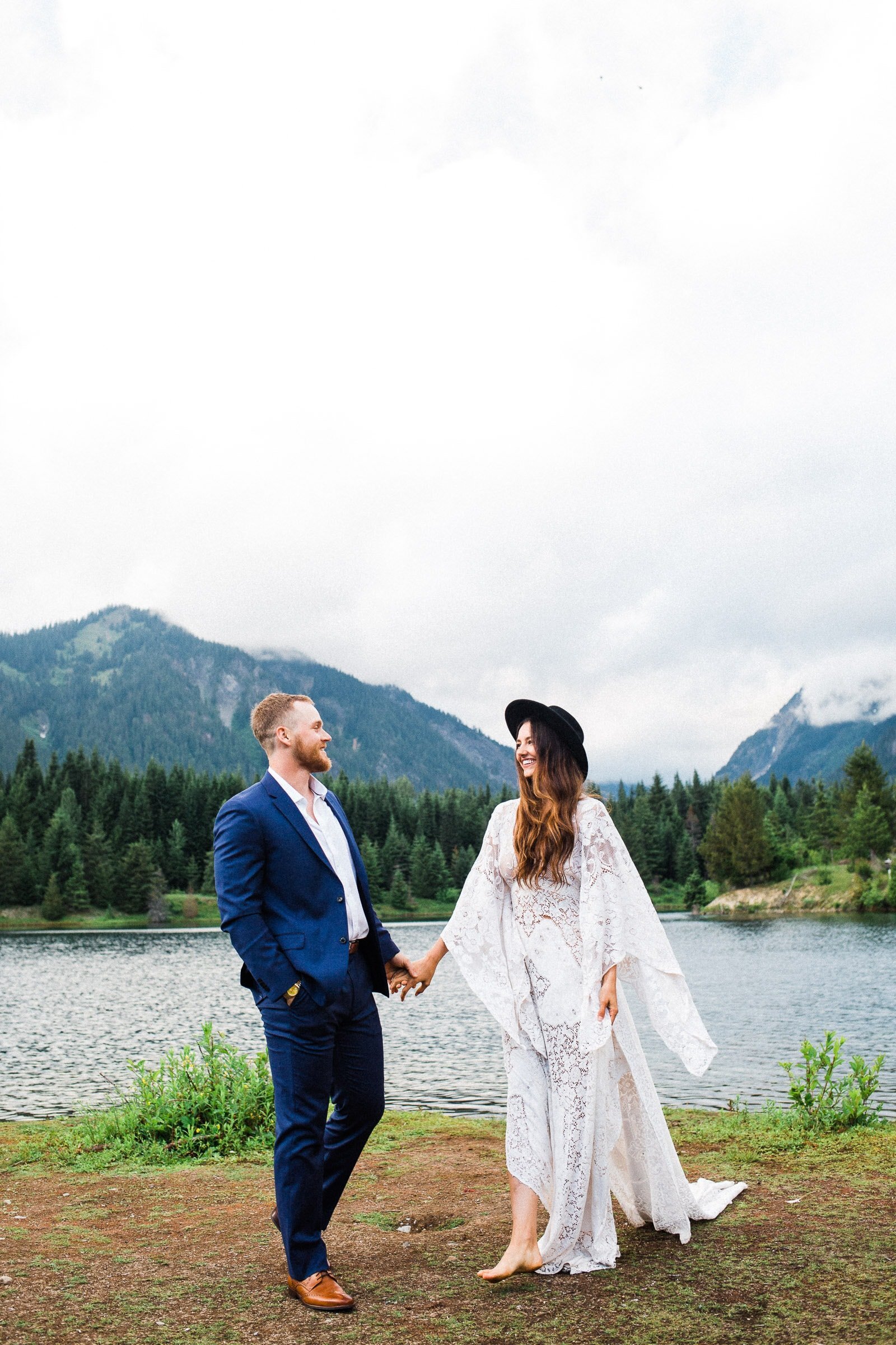 Couple holding hands and walking barefoot in front of mountains and lakeside in Washington State