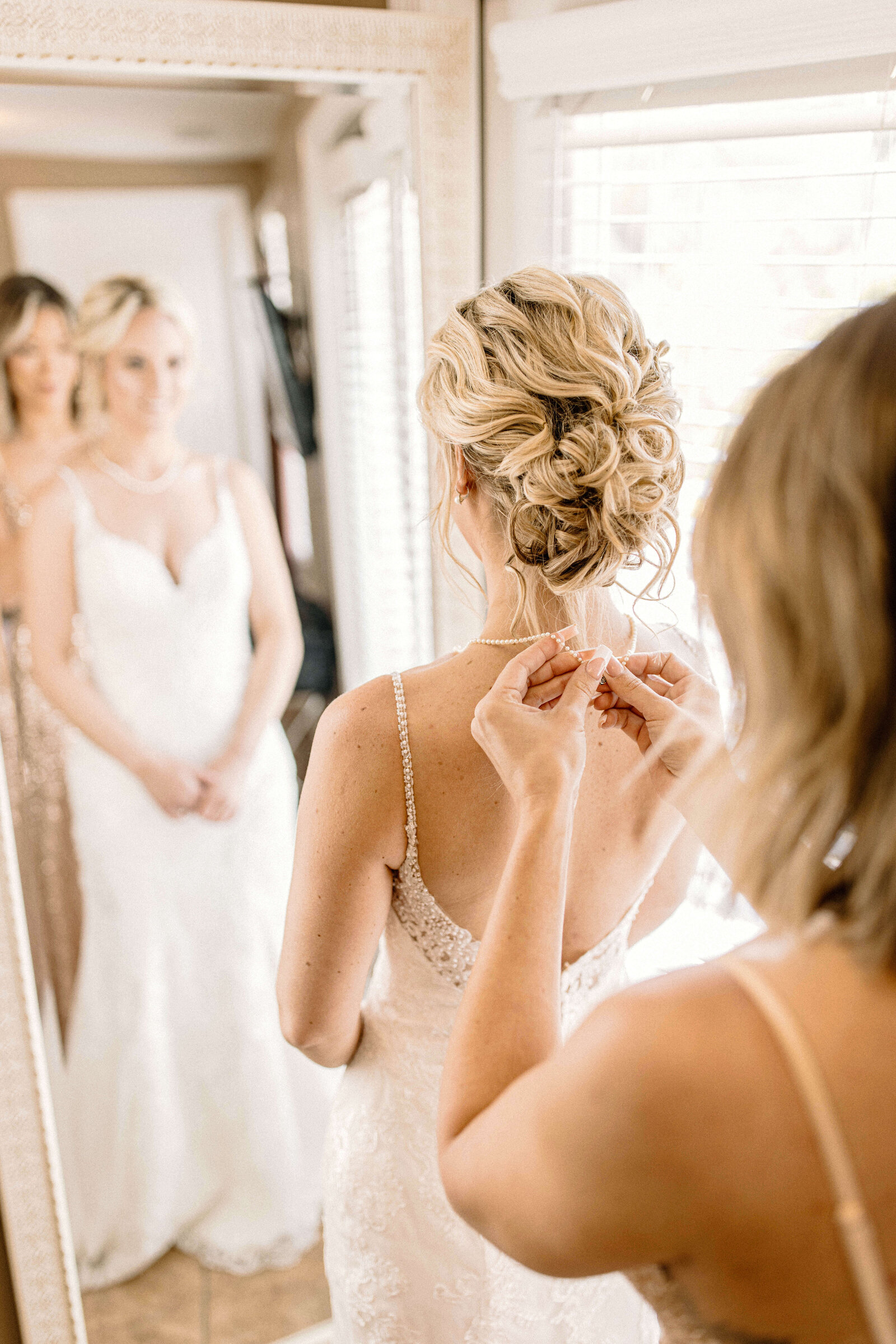 bridesmaid putting on brides necklace in getting ready suite by delaware wedding photographer sabrina leigh