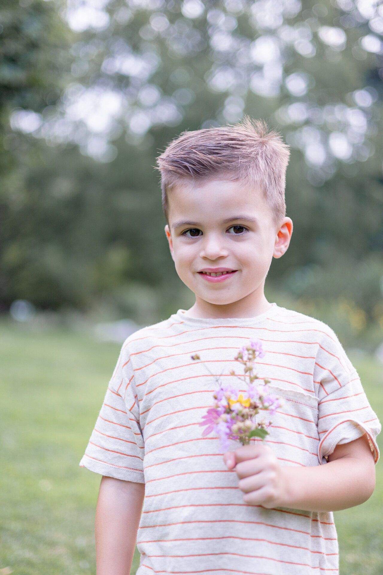 Young boy holding a bouquet of wildflowers and smiling softly during a summer outdoor portrait session in Fairfield County, CT. Dressed in a striped t-shirt with trees in the background, the photo captures a natural and candid moment.