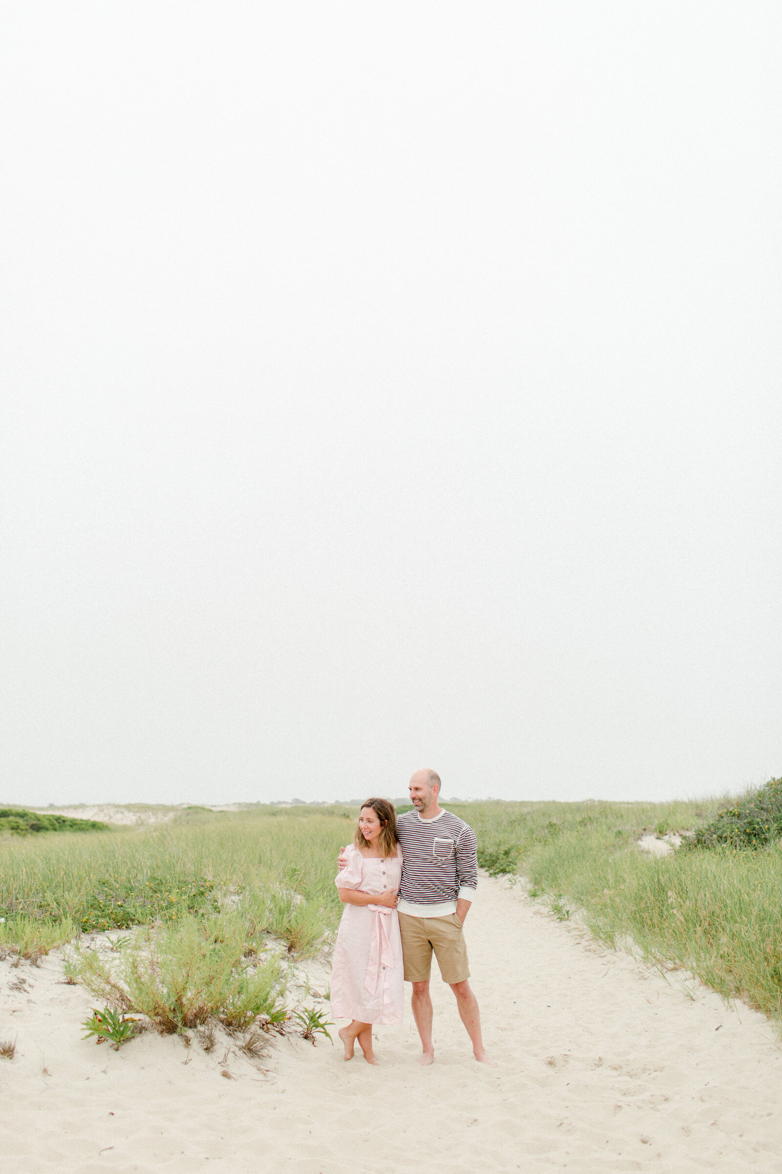 A husband and wife stand together on a sandy path at the beach while staring off to one side during photo session with Boston photographer Corinne Isabelle