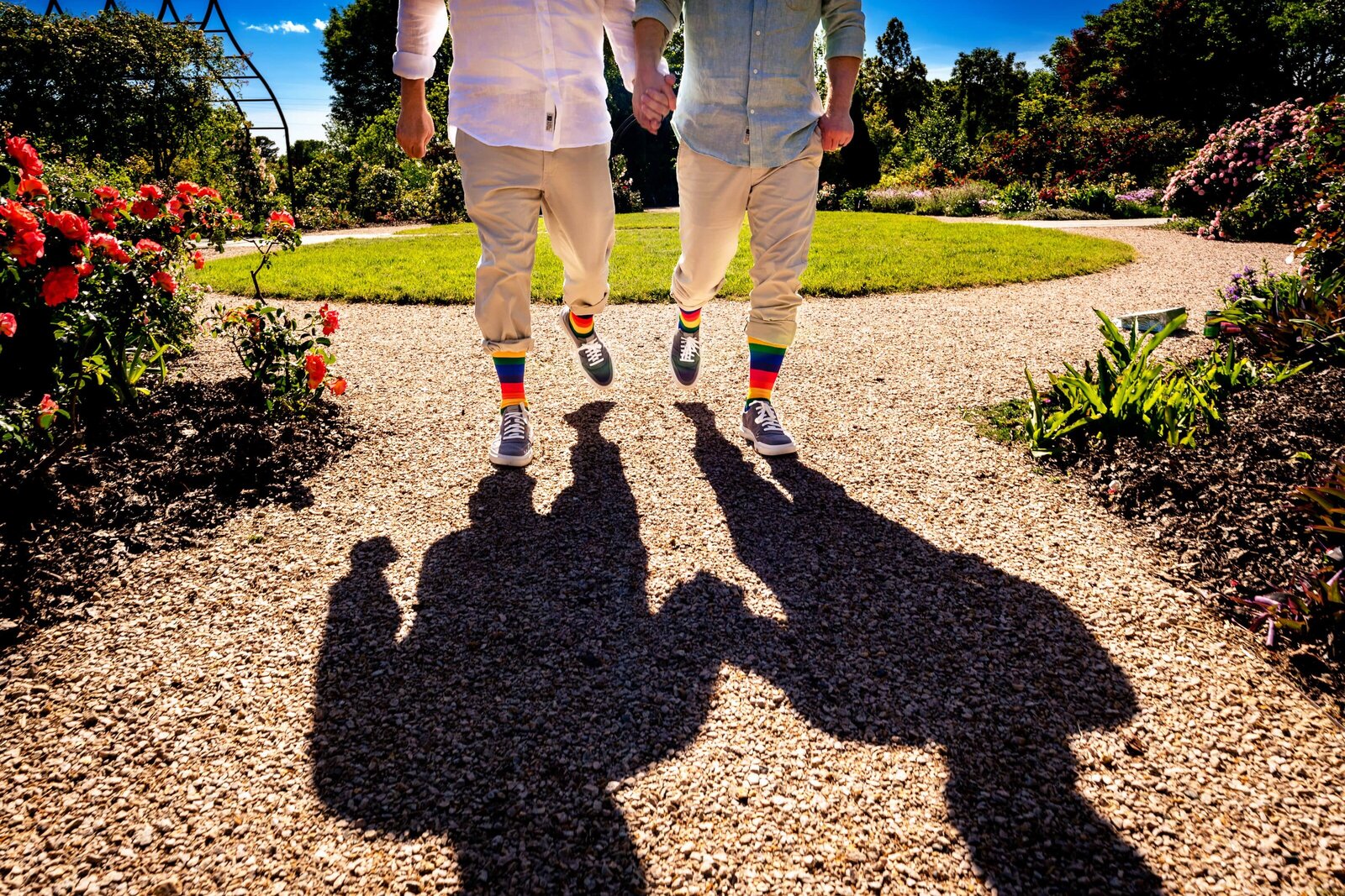 two men wearing coordinated outfits and rainbow socks holding hands and walking toward the camera. The frame shows them from chest down  with their shadows extending in front of them toward the camera