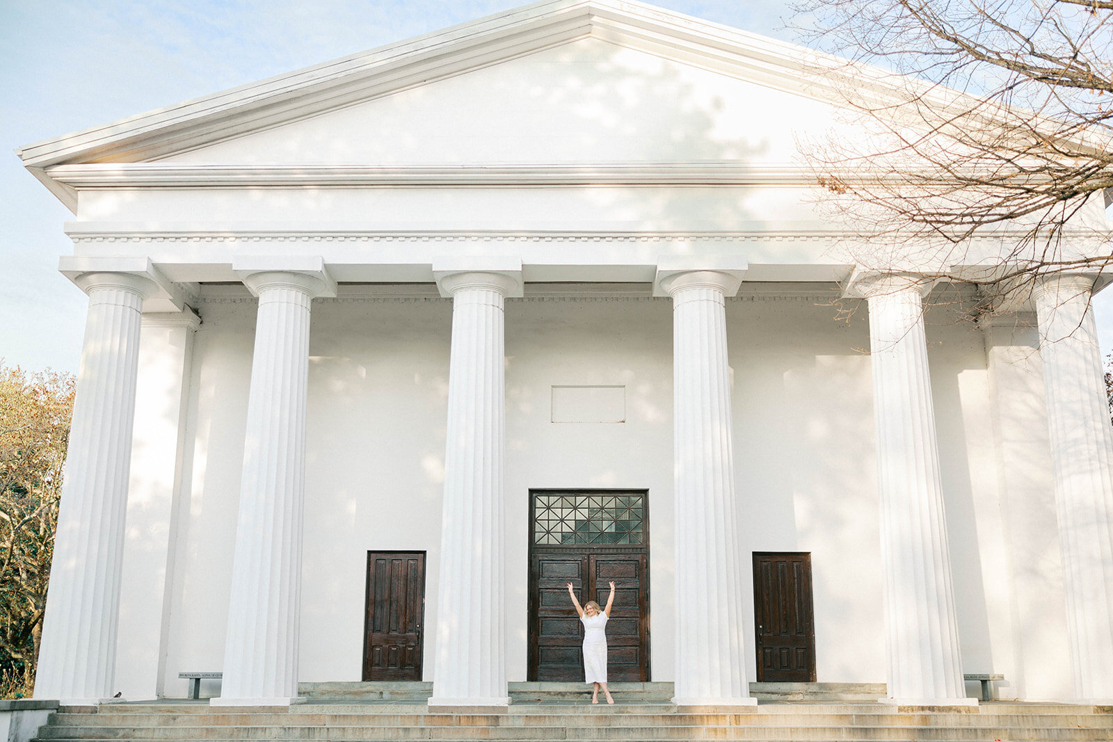 UGA Grad in cap and white dress raises arms up on the porch of The Chapel on North Campus