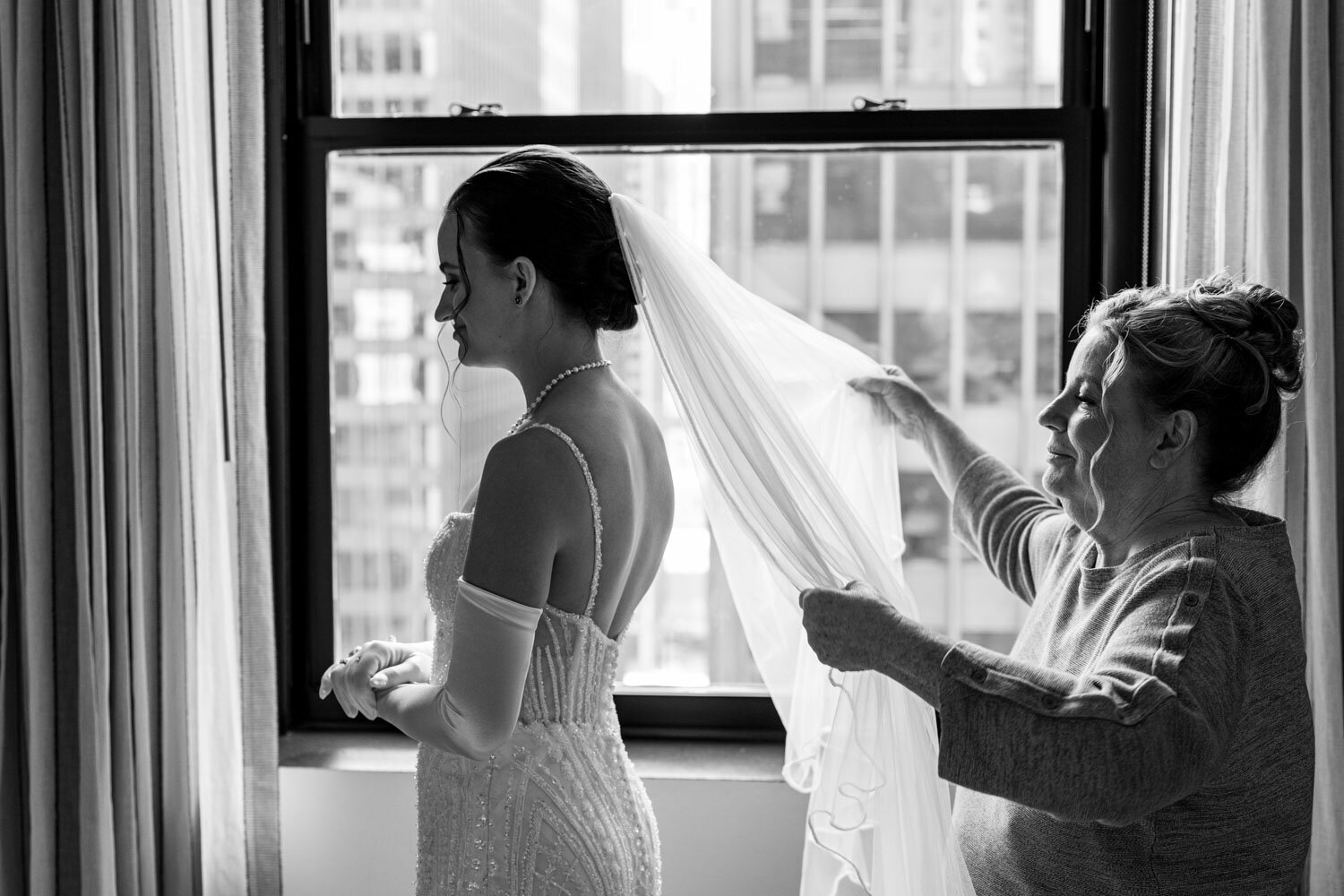 A woman is putting a veil on a bride in front of a window.
