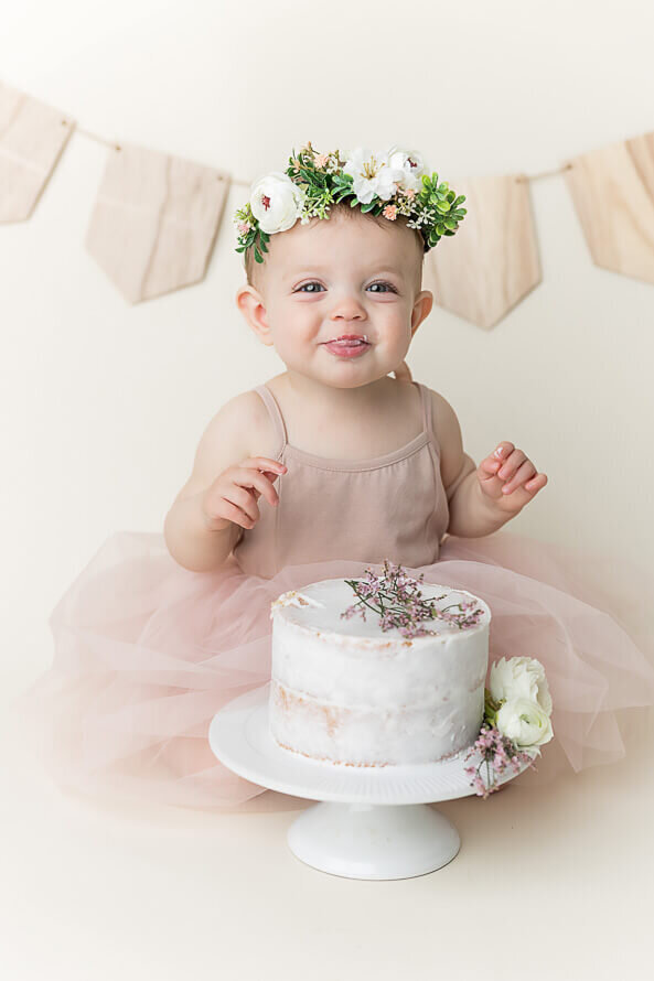 Baby in flower crown sticks her tongue out at camera with her birthday cake.