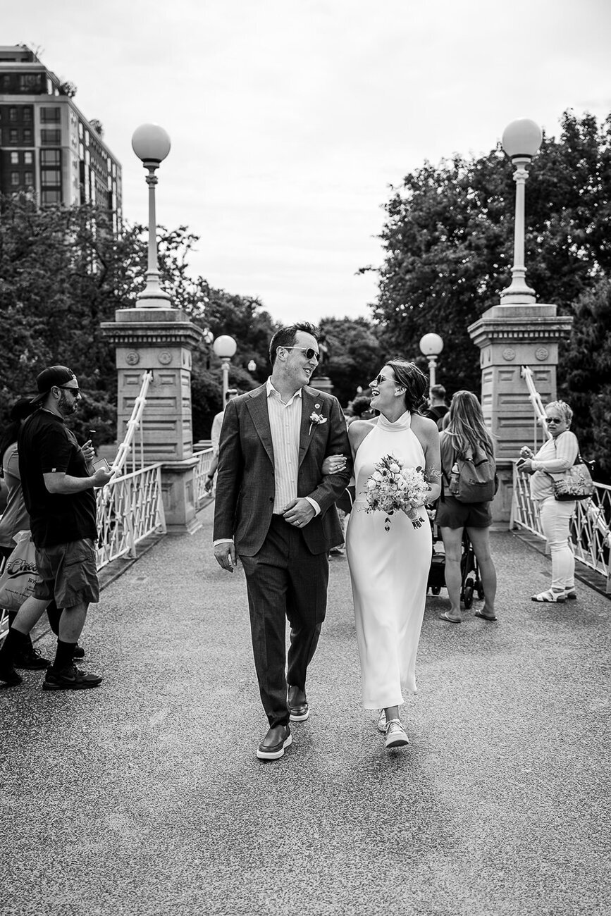 black and white portrait of bride and groom wearing sunglasses and walking across the bridge in the boston public garden