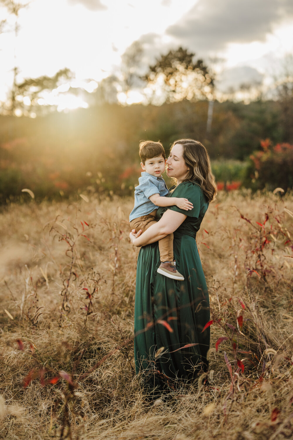 pregnant mother in green dress stand in field holding toddler boy at sunset