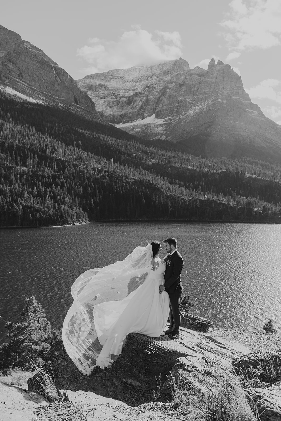 A black and white photo of a couple standing on a rocky ledge by a mountain lake, the bride’s veil blowing in the wind as they share an intimate moment during their Glacier National Park elopement, captured by Sydney Breann Photography.
