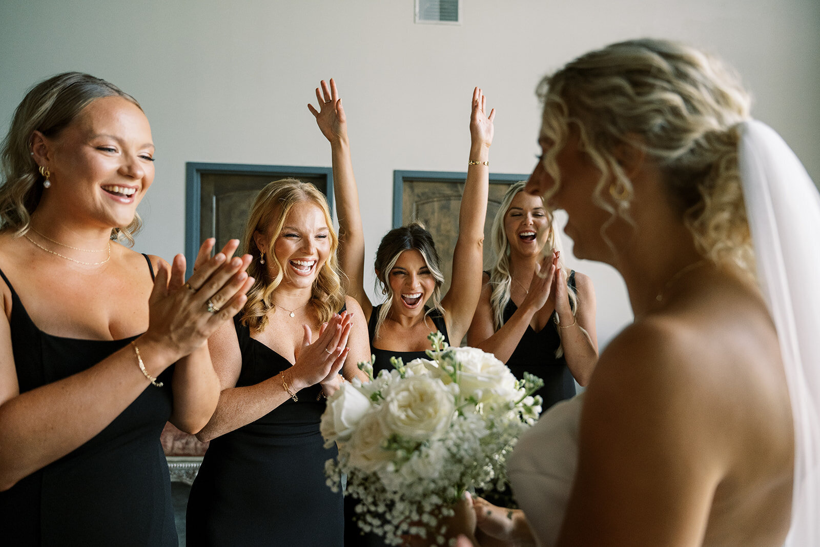 Bride shares an emotional moment with her bridesmaids, who cheer and clap in excitement as she holds her bouquet. A candid, joyful wedding morning captured in a documentary photography style.