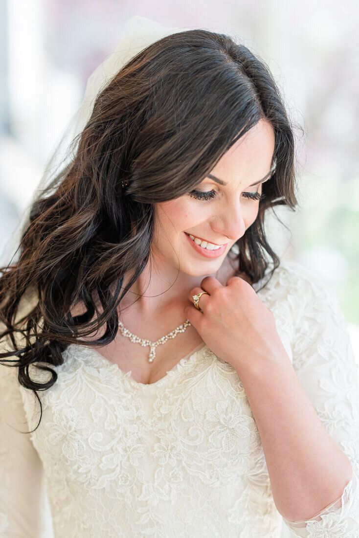 A bride wearing a cream colored lace wedding dress smiles softly and looks down as she stands in front of a window, preparing for her spring wedding in Pleasant Grove.