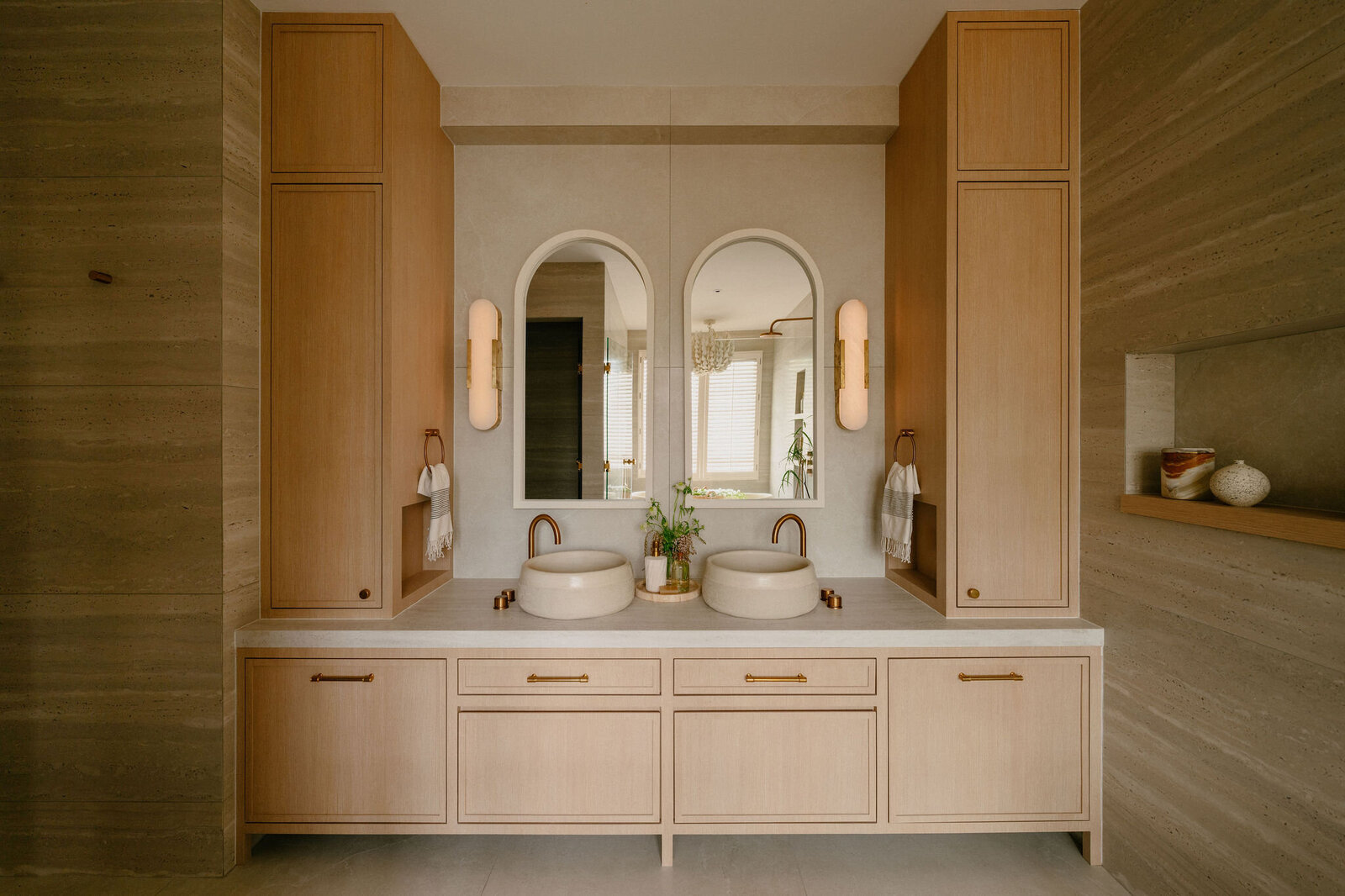 Bathroom vanity with twin stone basins, arched mirrors, and brushed brass tapware framed by oak cabinetry.