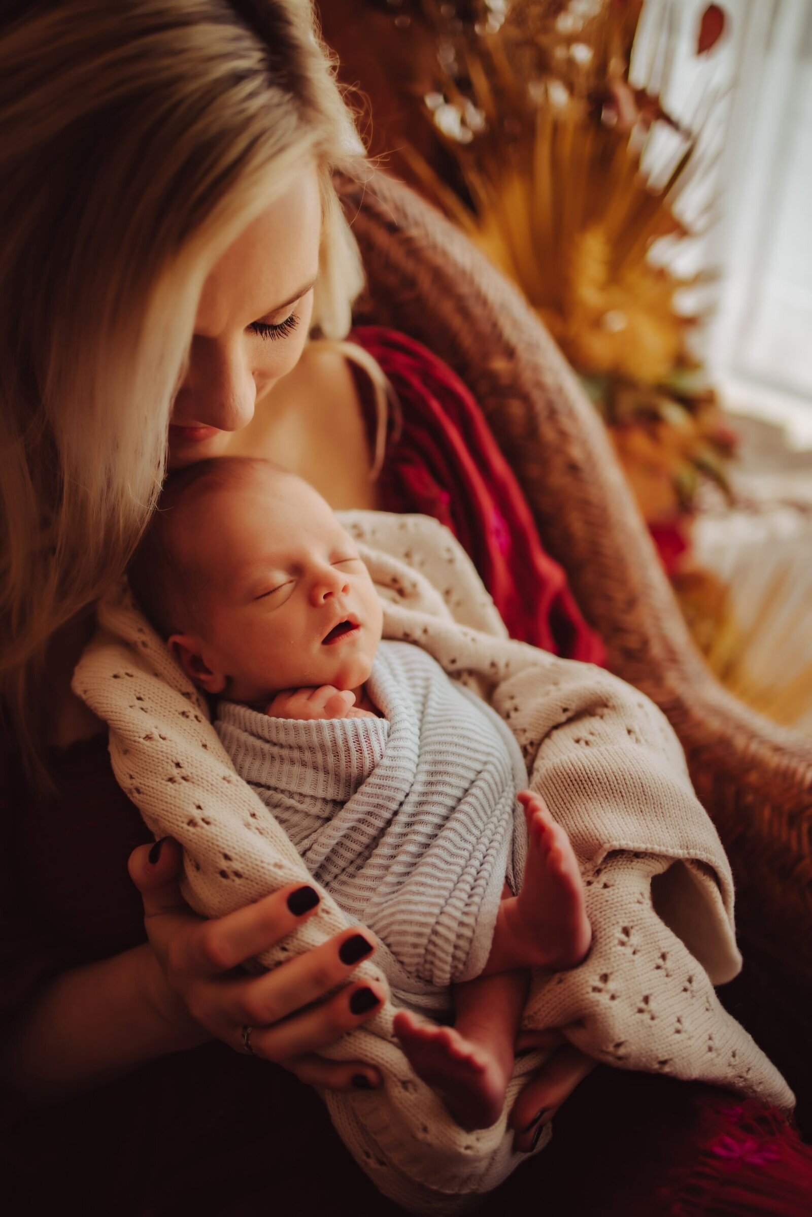 A woman sitting in a chair, holding her swaddled newborn baby as she  plants a kiss on their forehead captured by Infinite Productions