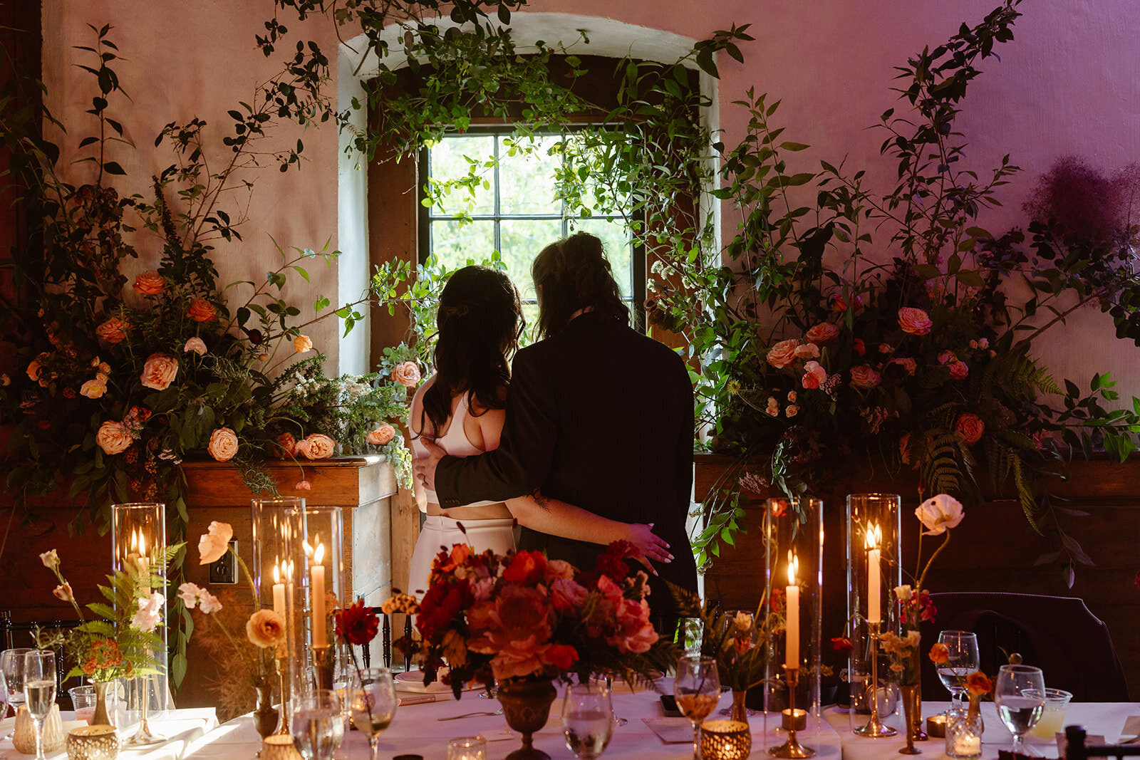 A couple stands looking out of a window. They are surrounded by candles and plants like they are apart of the secret garden.