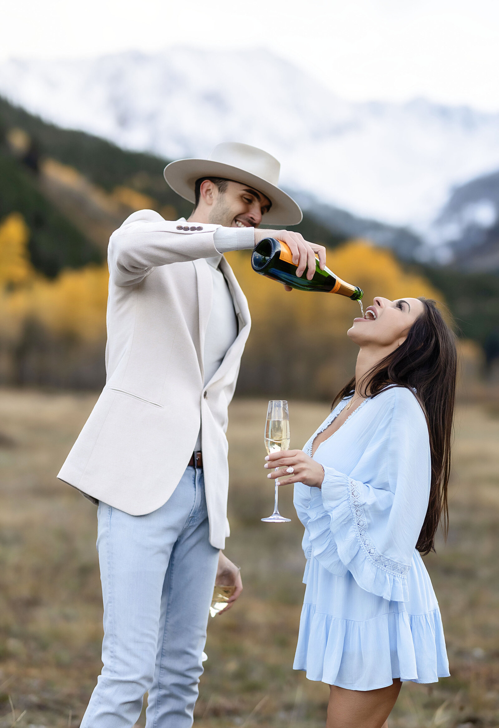 A man pours some champagne into his fiances mouth as they celebrate their Aspen mountain engagement.