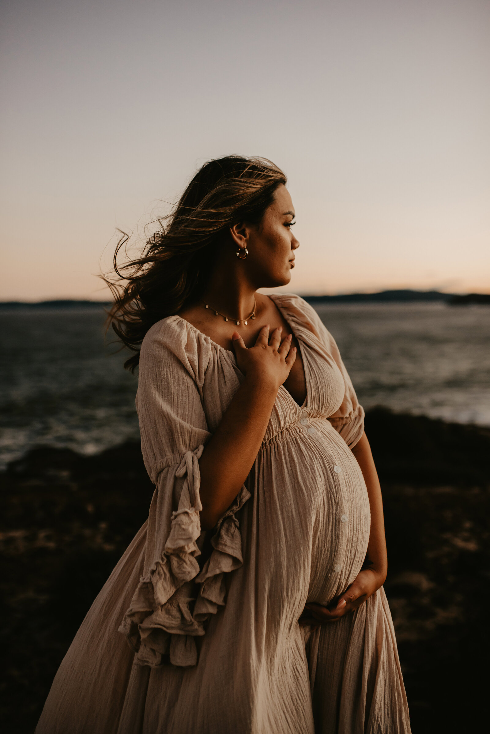 A pregnant woman in a flowing dress stands by the seaside at dusk, looking into the distance with her hand on her chest.