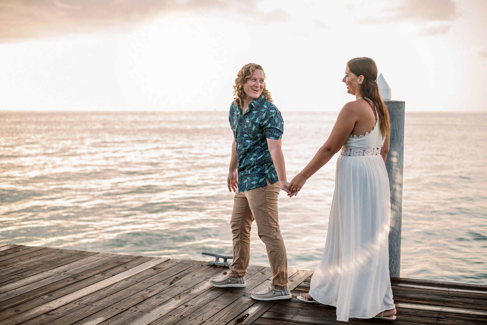 engaged lesbian couple walking on a boardwalk together as the sun is setting. One woman is leading the other while looking back at her and smiling