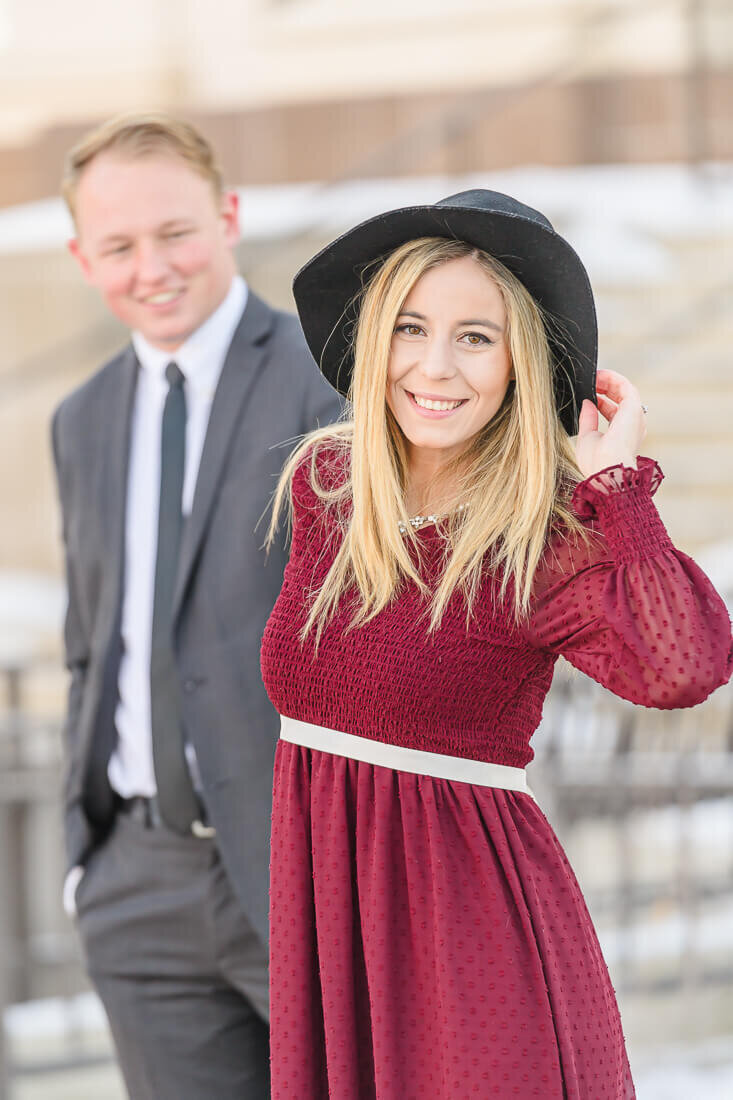 A woman in a long sleeve burgundy dress walking towards the camera, pulling her fiance in a gray suit along as they walk on the grounds of BYU, Provo. Captured by Utah engagement photographer Melissa Woodruff Photography