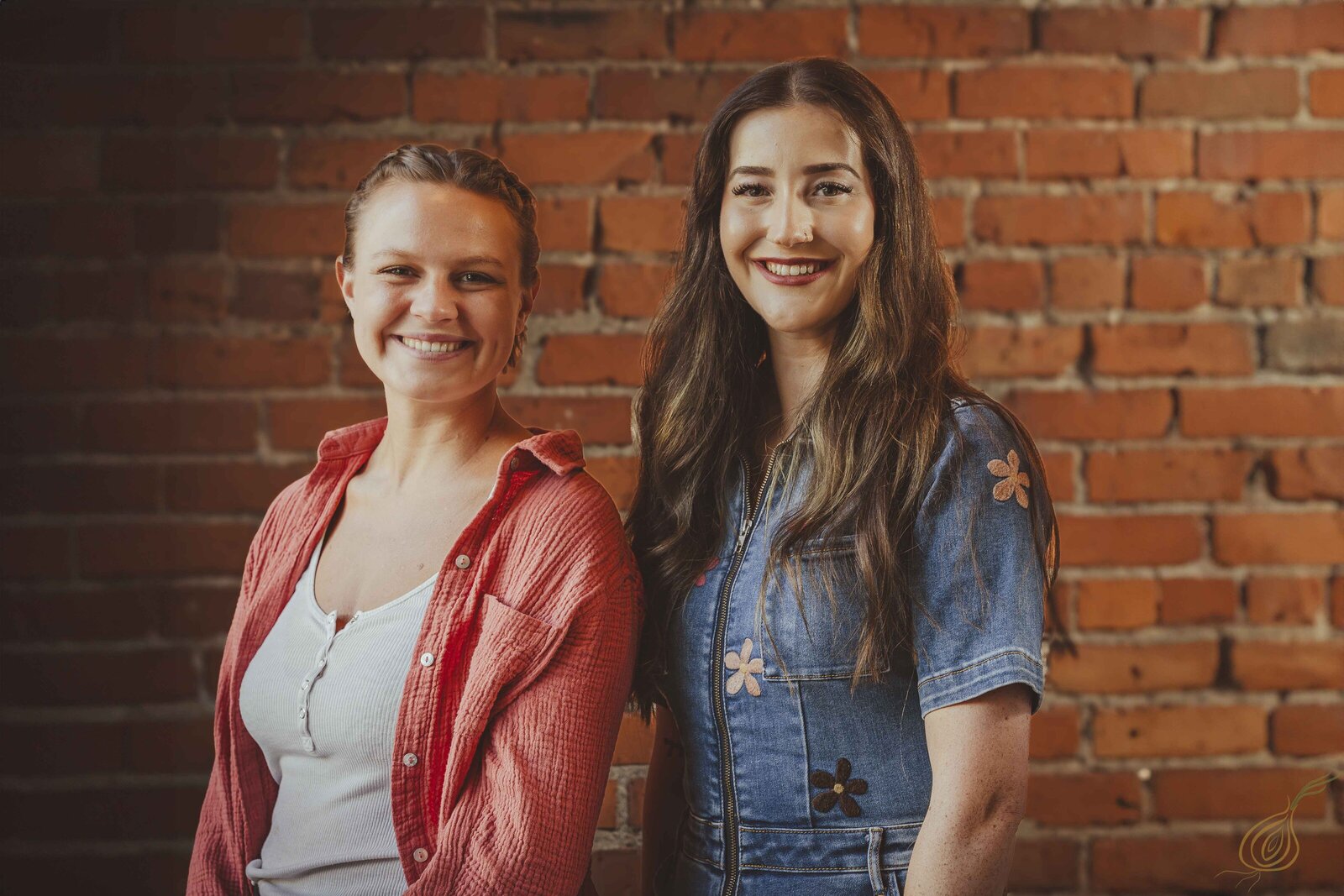 Two women stand side by side, smiling, in front of a textured brick wall, showcasing a casual and friendly atmosphere.
