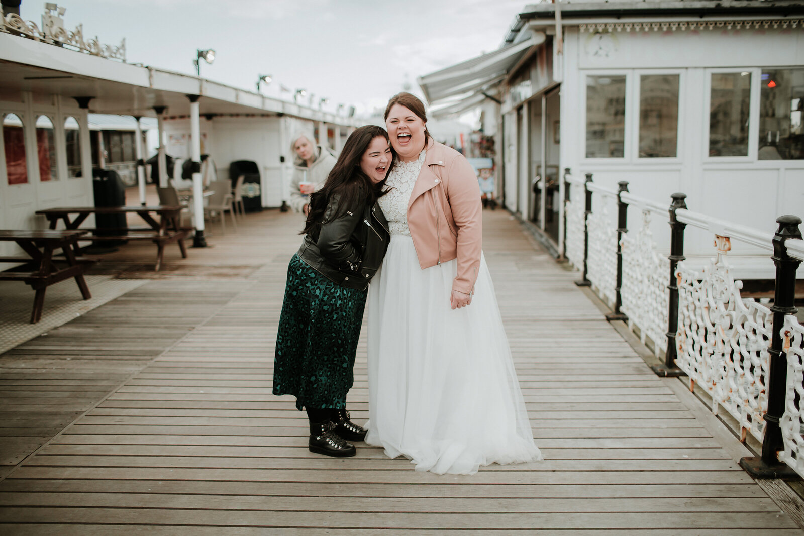 Bride in pink leather jacket with best friend laughing on Brighton Pier