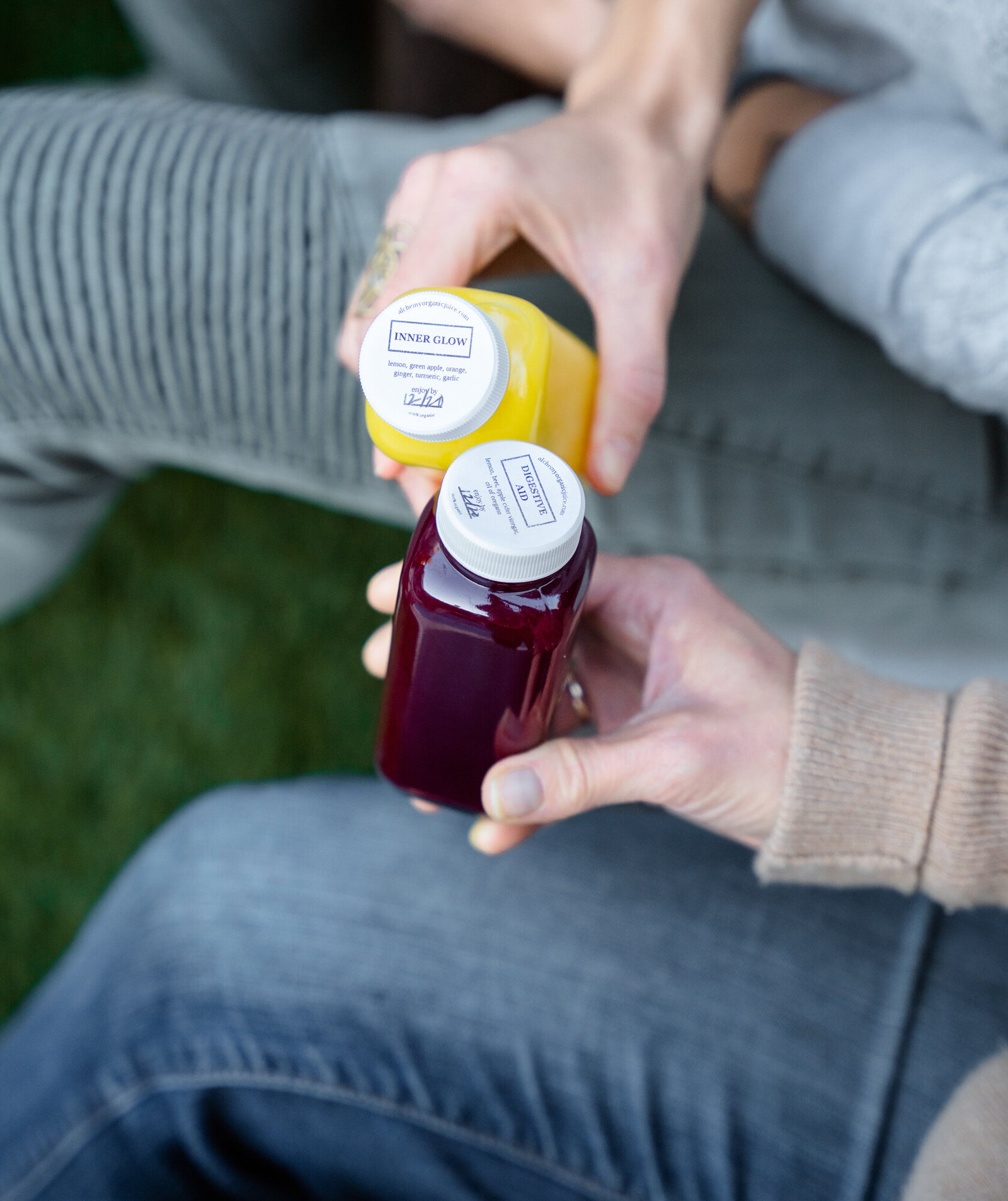 Branding Photographer, friends sit beside each other in the grass, they're holding juice bottles