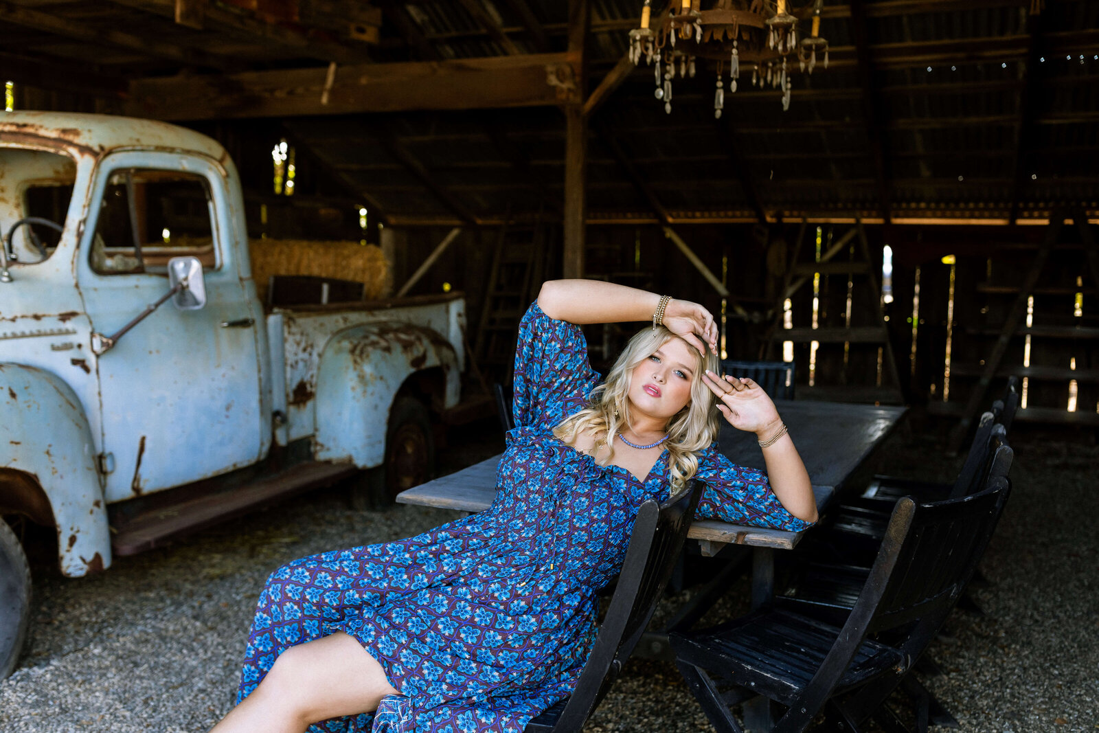 blonde girl wearing blue dress posing under a barn next to a rusted car and a black wagon
