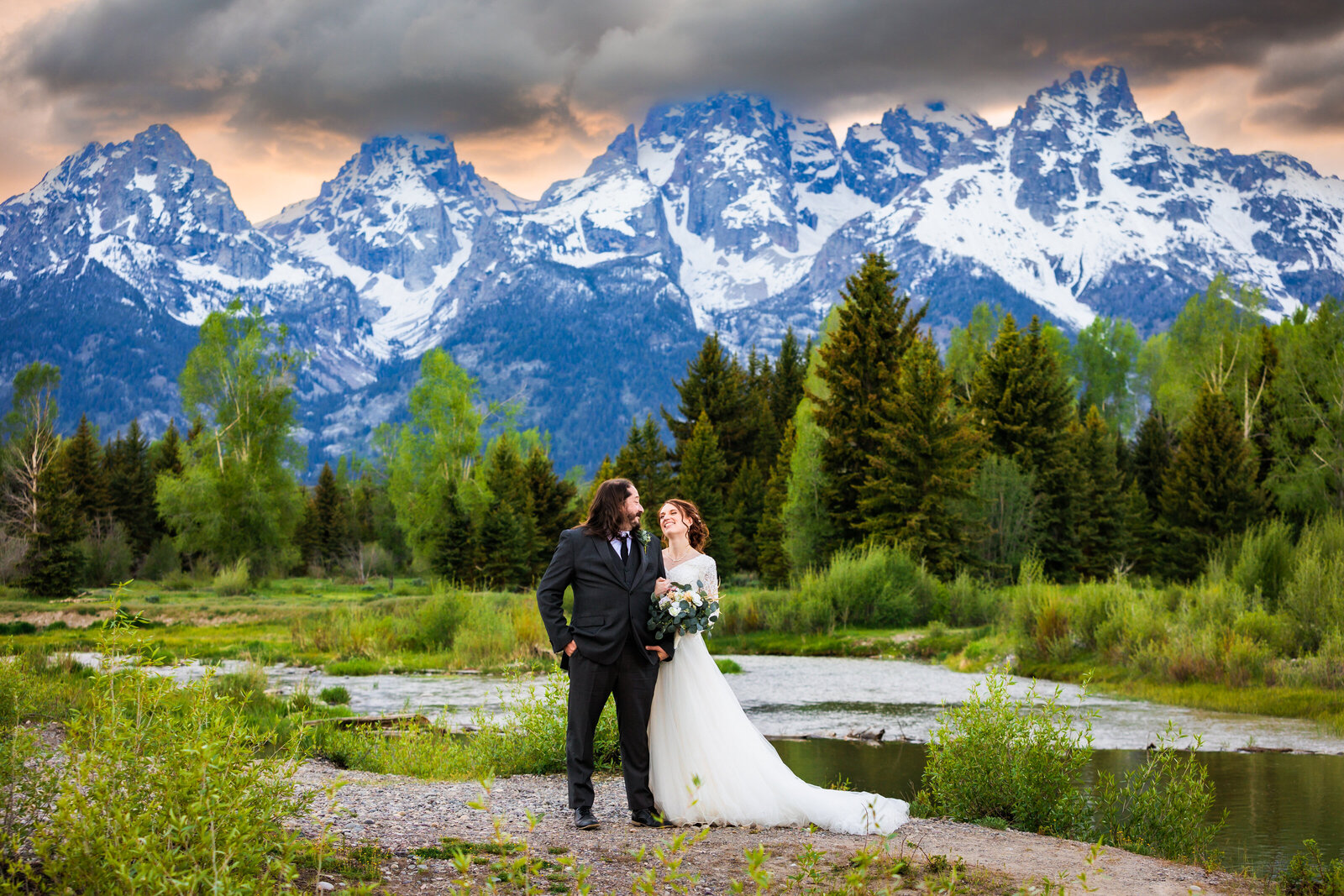 Grand Teton elopement photographer captures bride and groom standing in front of mountains with sunset behind them that is orange