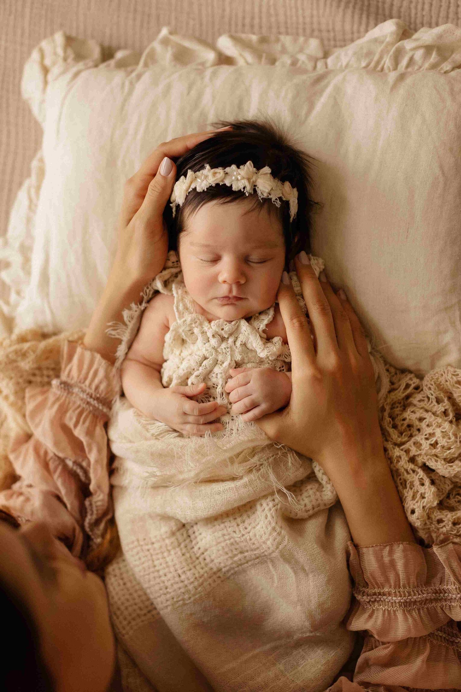 Newborn baby with dark hair and floral headband sleeping, parent's hands gently cradling head, soft beige fabrics