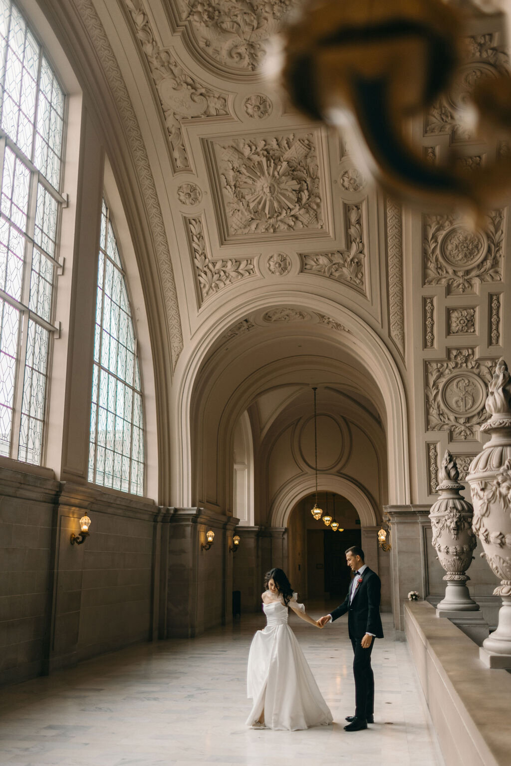 A bride and groom holding hands in a grand hall with ornate ceilings and large windows in San Francisco City Hall, showcasing intricate architectural details.