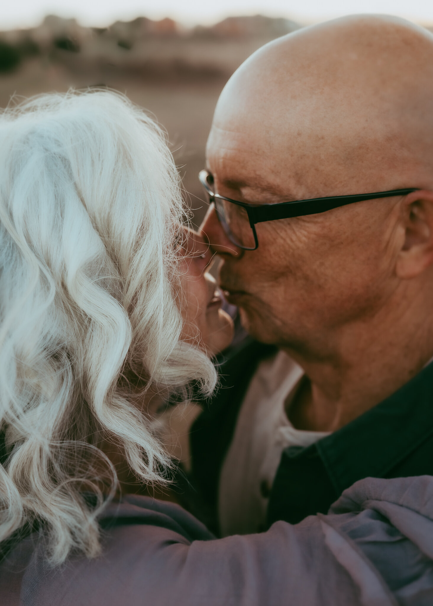 An older husband and wife kiss, while her bright white hair stand out in contrast to his bald head.