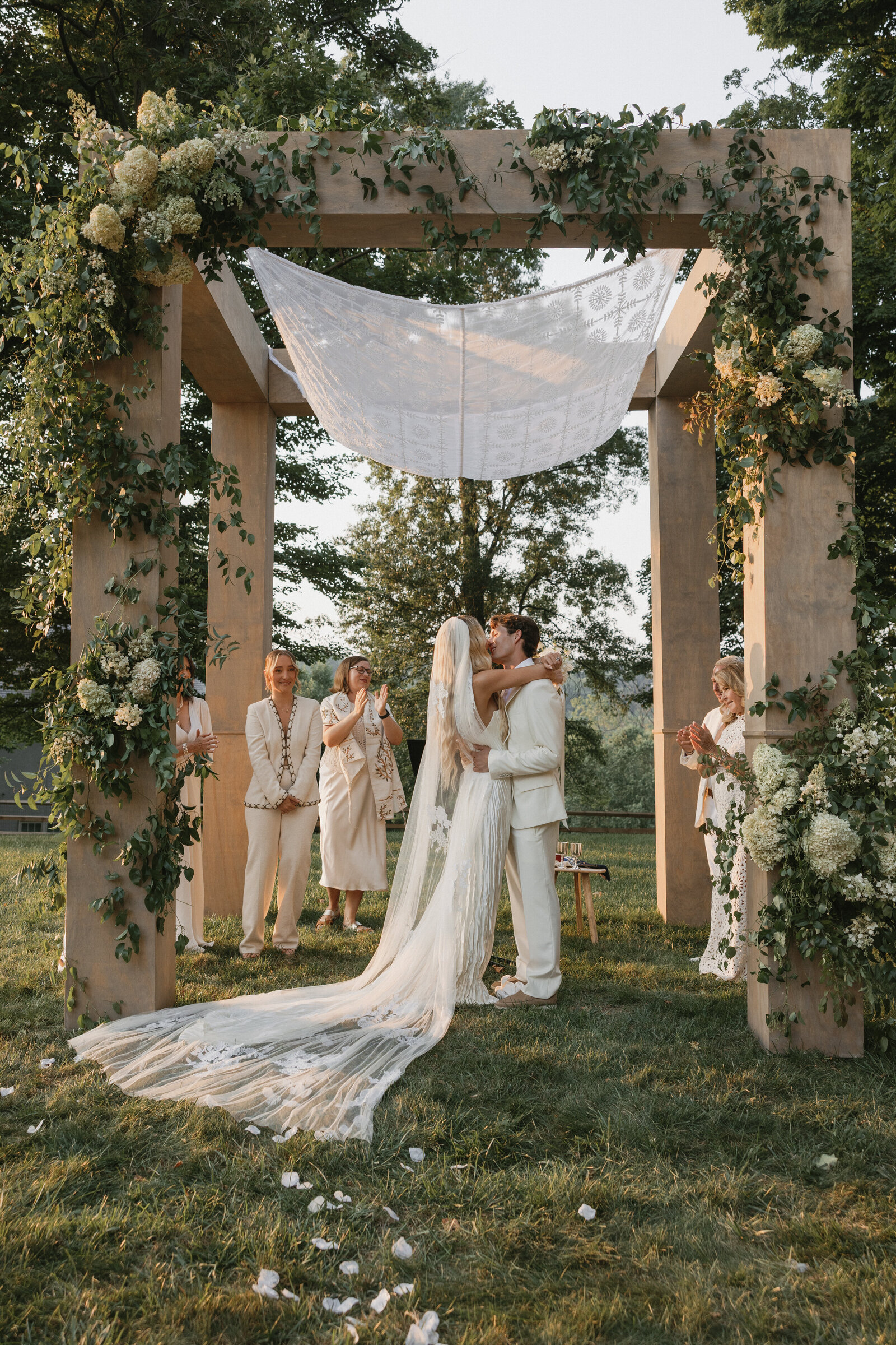 Bride and groom dressed in all white having first kiss under large wooden chuppah draped in white and green flowers at golden hour 