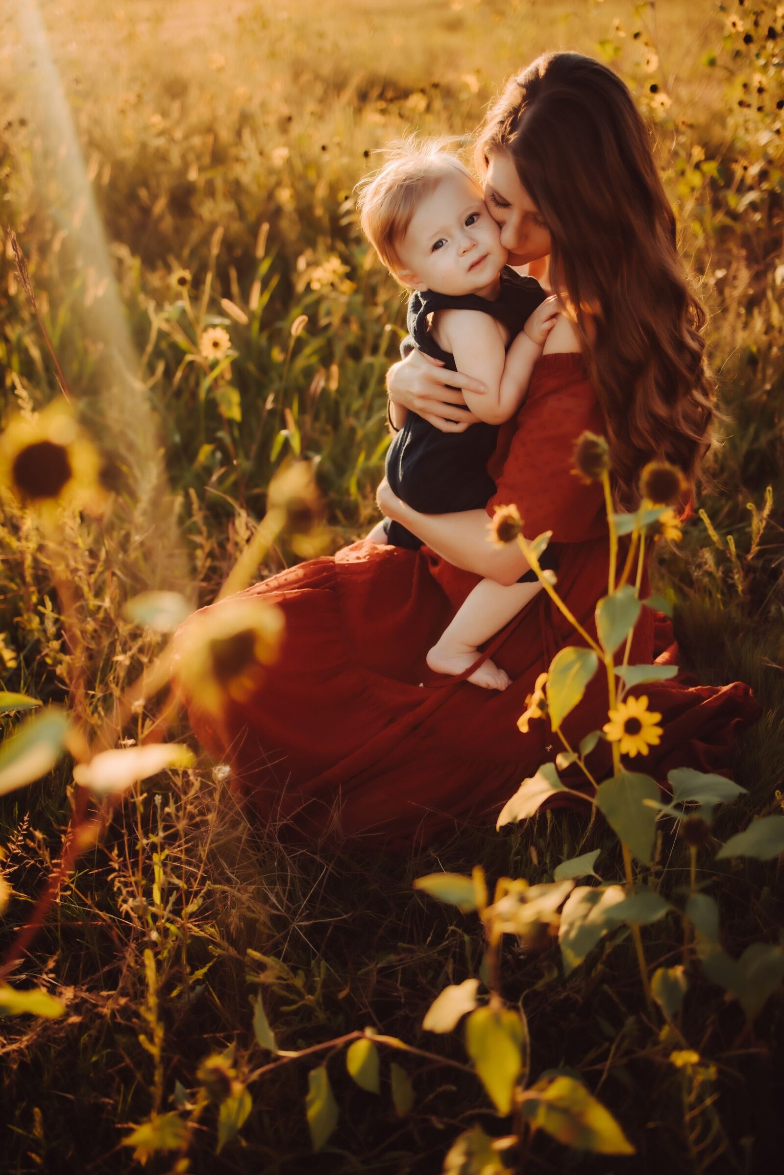 A woman in a red shirt holding her son close and grazing her lips against his cheeks while he looks into the camera during their mother/son family photoshoot