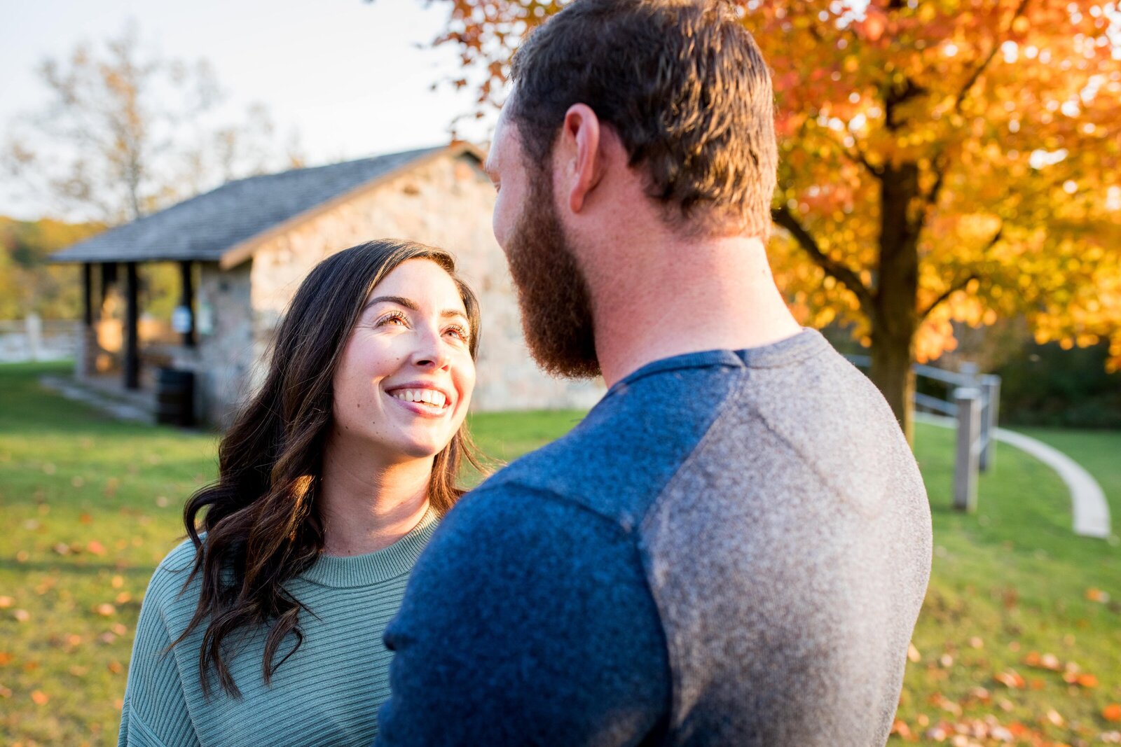 Woman looks at fiance during engagement session with fall trees in background.