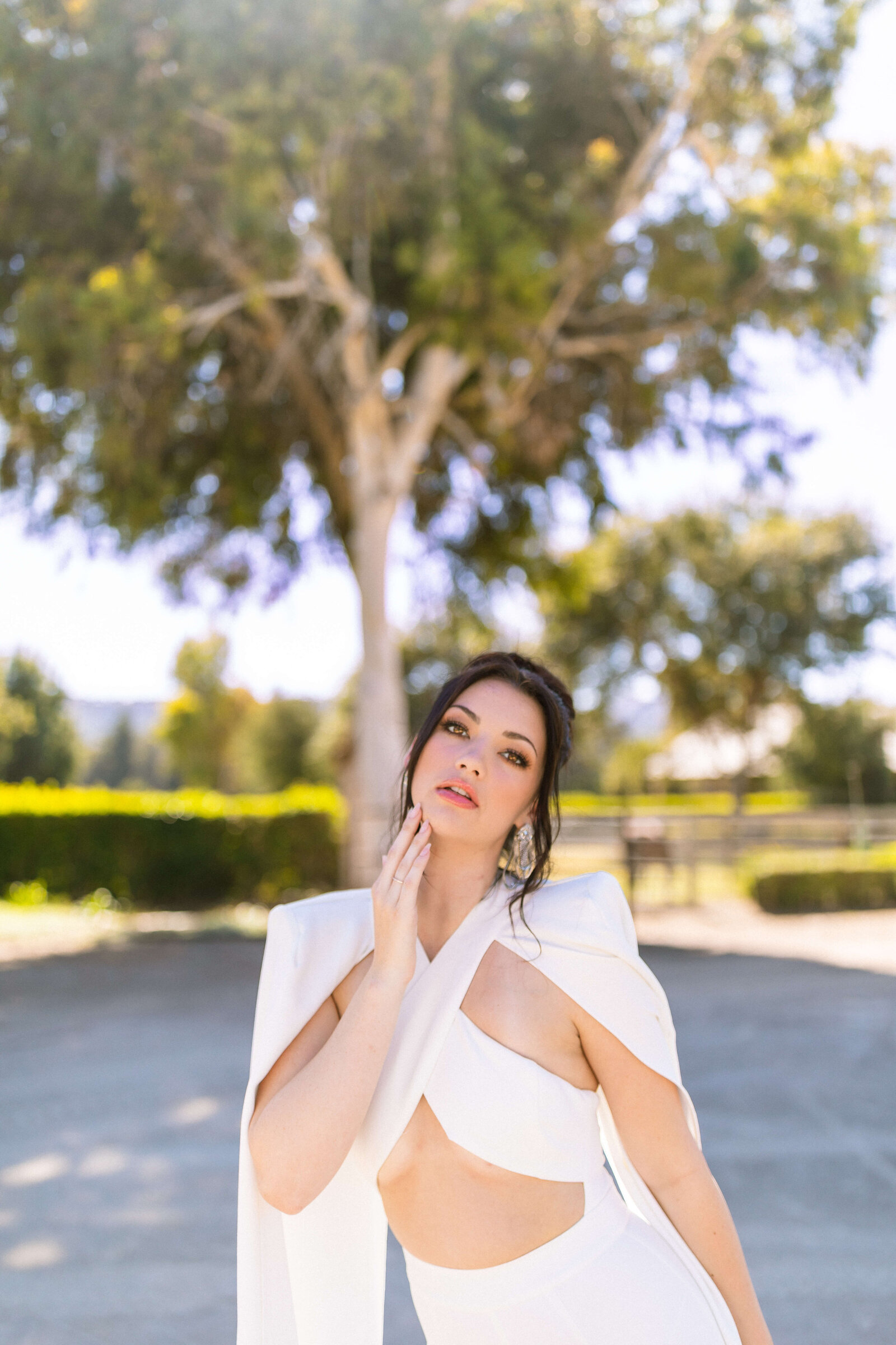 girl with dark hair in an updo wearing a white outfit posing for camera in front of a green tree