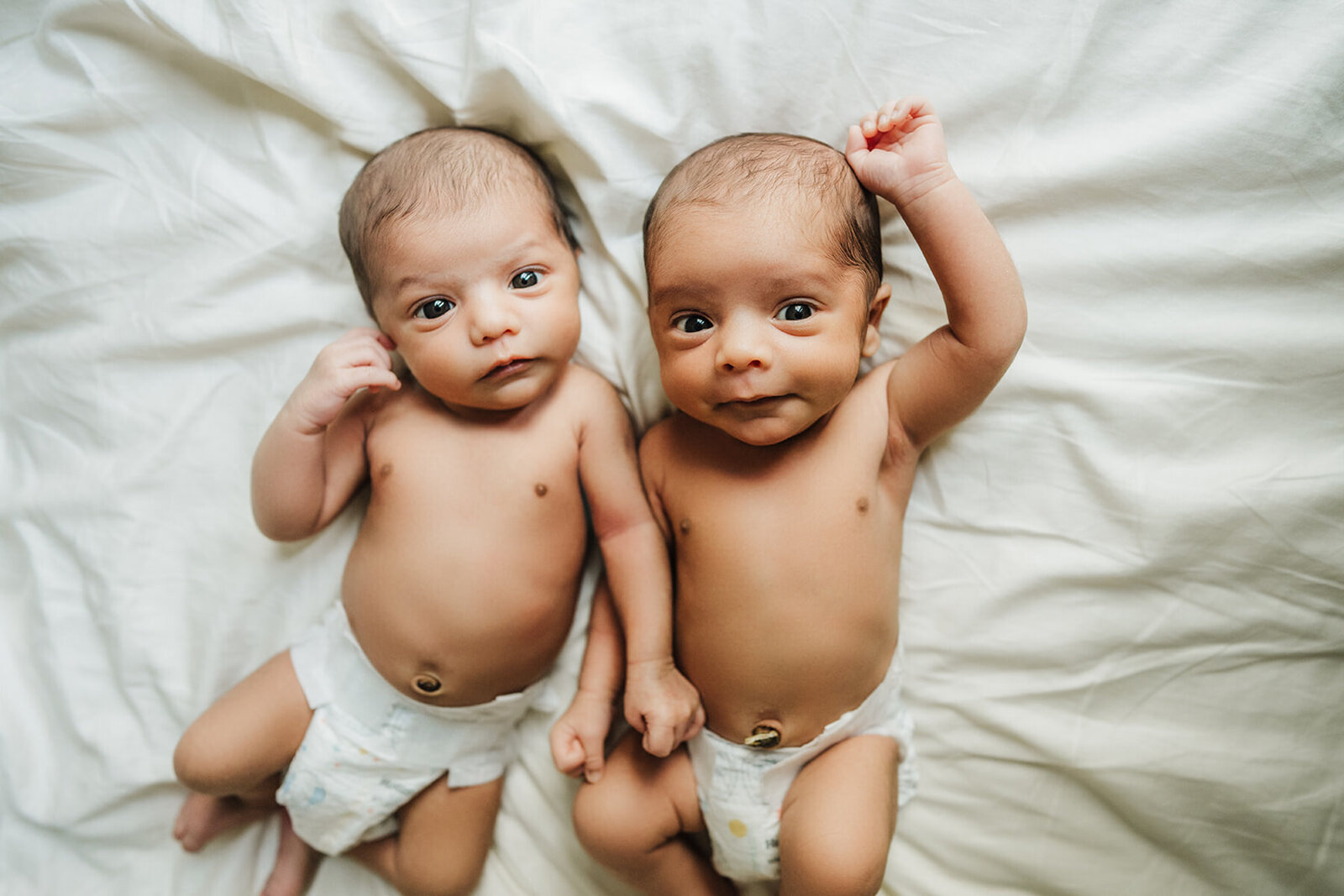 twin babies in diapers lay on white bedspread and stare at camera