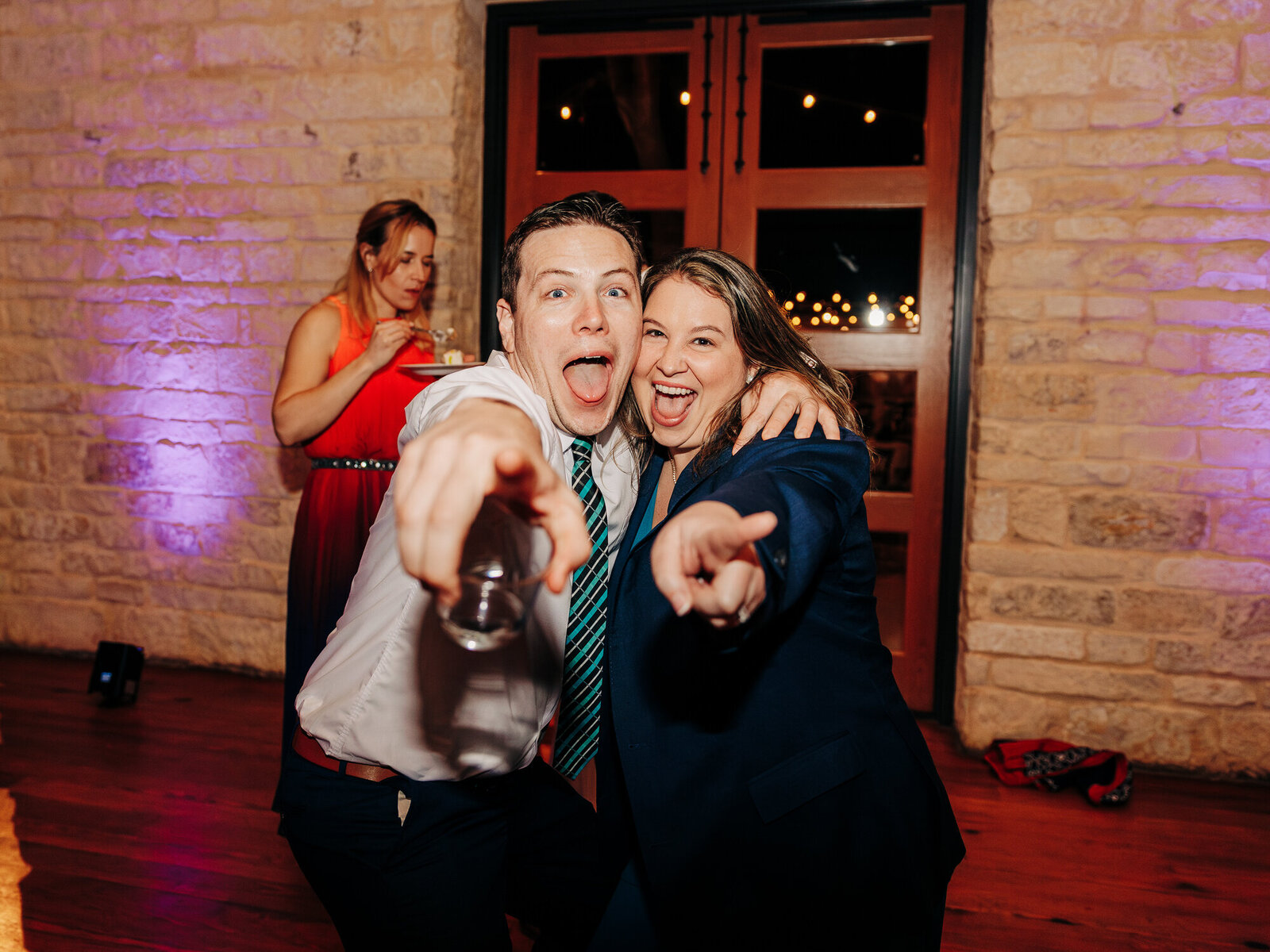 Two people in formal attire smile and enthusiastically point at the camera, standing close together at an indoor event with stone walls—captured perfectly by this Eau Claire wedding photographer. In the background, a guest in a red dress holds a plate of food.