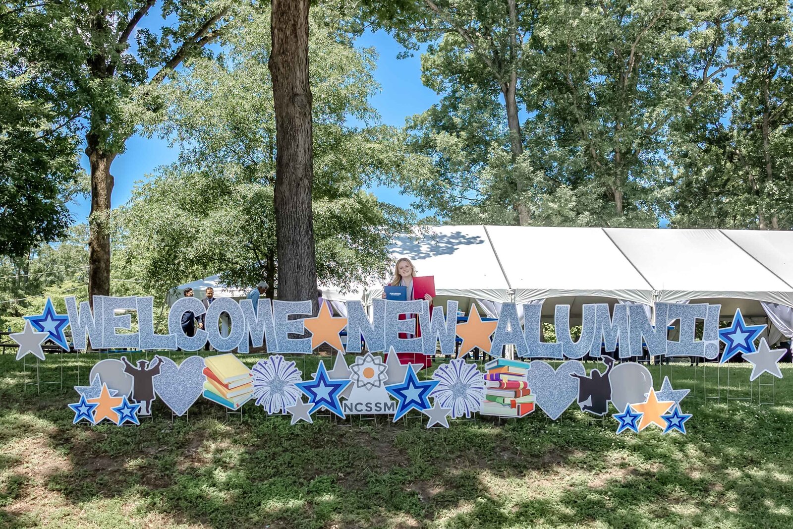 A decorative "Welcome New Alumni" sign set up in a park with trees and tents in the background at the North Carolina School of Math and Science in Durham, NC. This celebratory scene captures the spirit of graduation events, perfect for alumni gatherings and school photography.