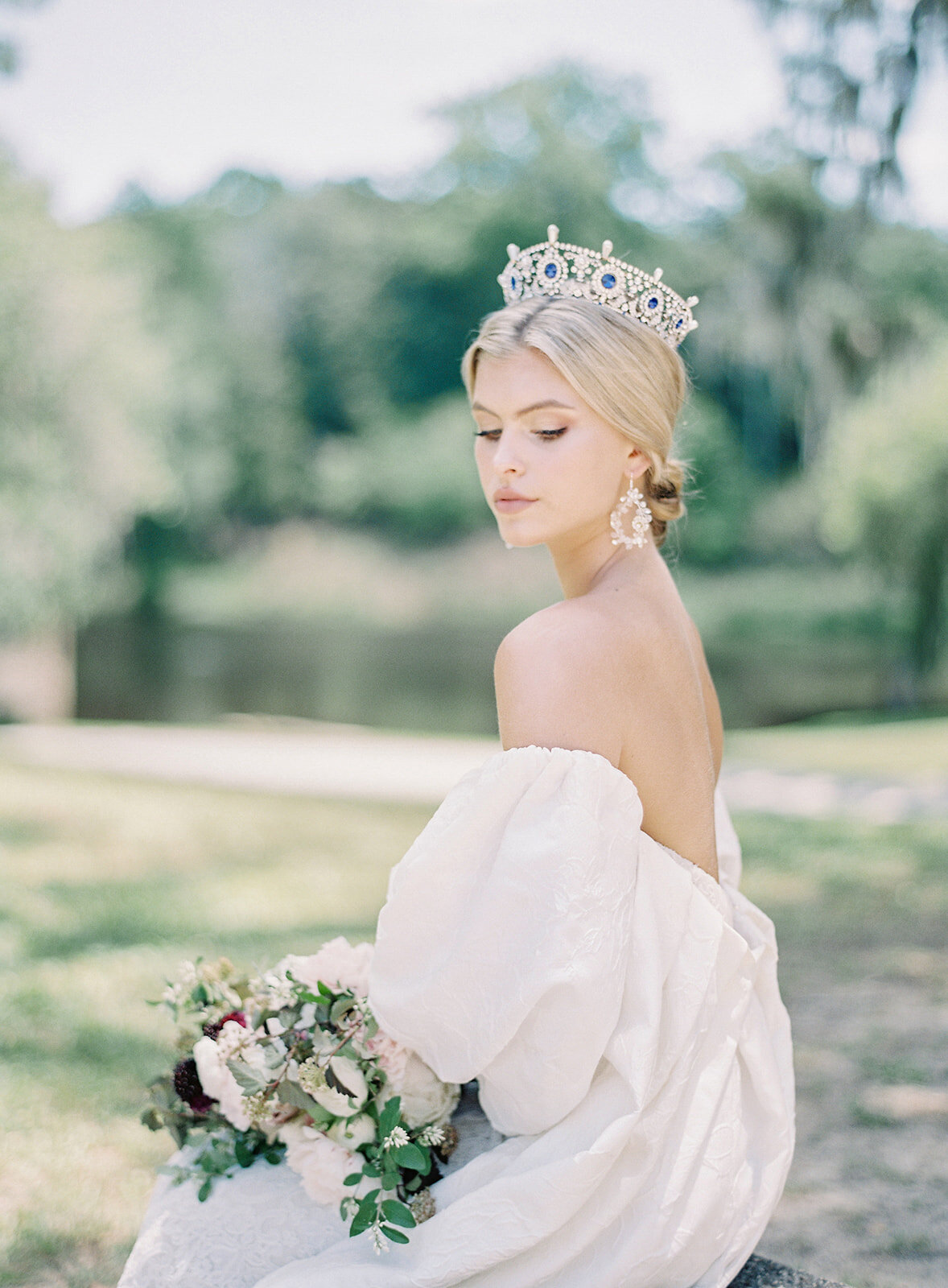 Bride wearing a sapphire crown with her hair in a low chignon bun and drop earrings. She is wearing a strapless wedding gown and cape that hangs from her upper arm so that her shoulders and back are showing. She is looking down at her bouquet on her lap. In the background are oak trees and water. Photographed by wedding photographers in Charleston Amy Mulder Photography.