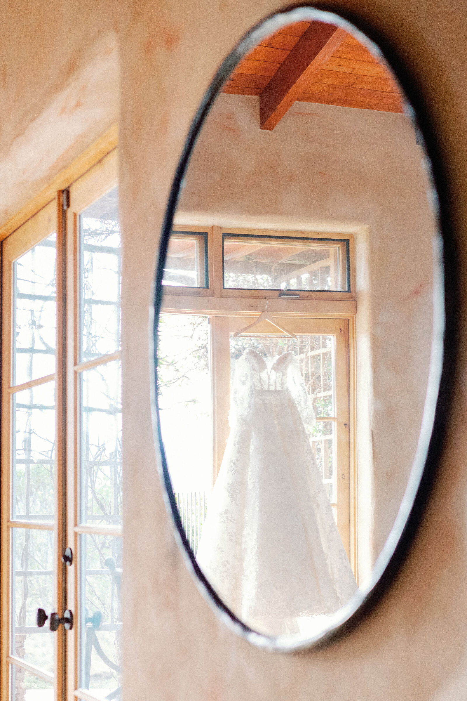 wedding photographer bay area captures detail shot of bridal gown hanging in a door with sunshine coming in through the windows reflected in a circle mirror