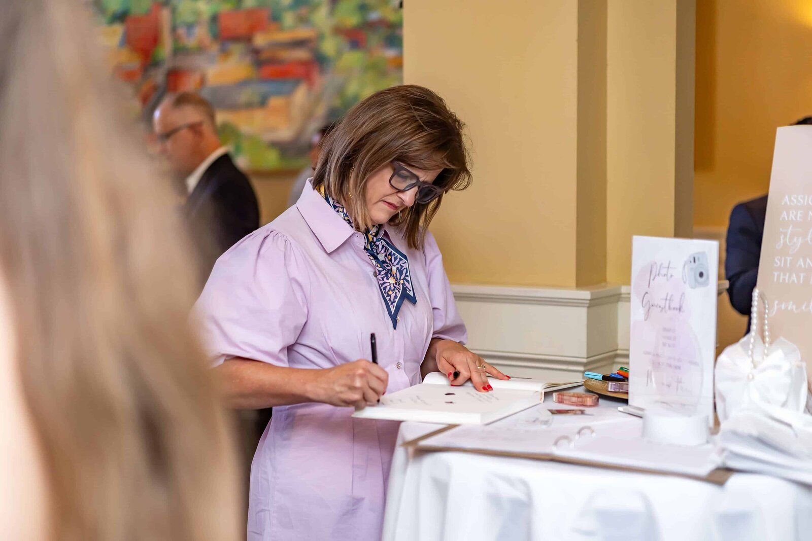 A woman wearing glasses and a light purple dress signs a book at a standing table during an indoor event. Other people and colorful artwork are visible in the blurred background.