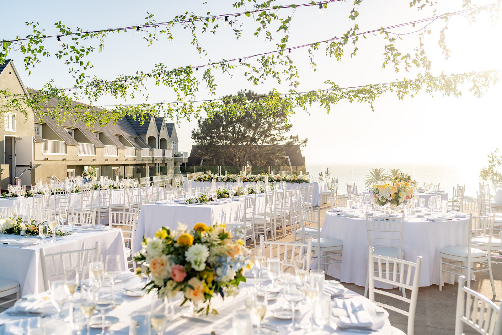 A wide shot of an outdoor reception decoration with white linen tables, colorful floral center pieces, and greenery hanging above