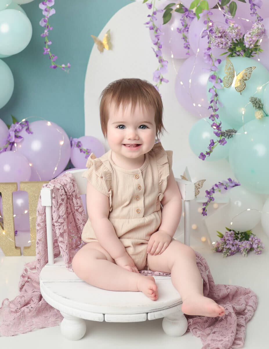 A charming girl sitting in a white chair, surrounded by vibrant purple and teal balloons, with a beautifully crafted lilac-themed cake smash setup in a studio in Rochester, NY.