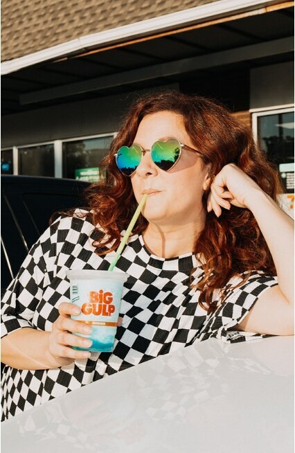 A woman leaning on a car sipping a slurpee.