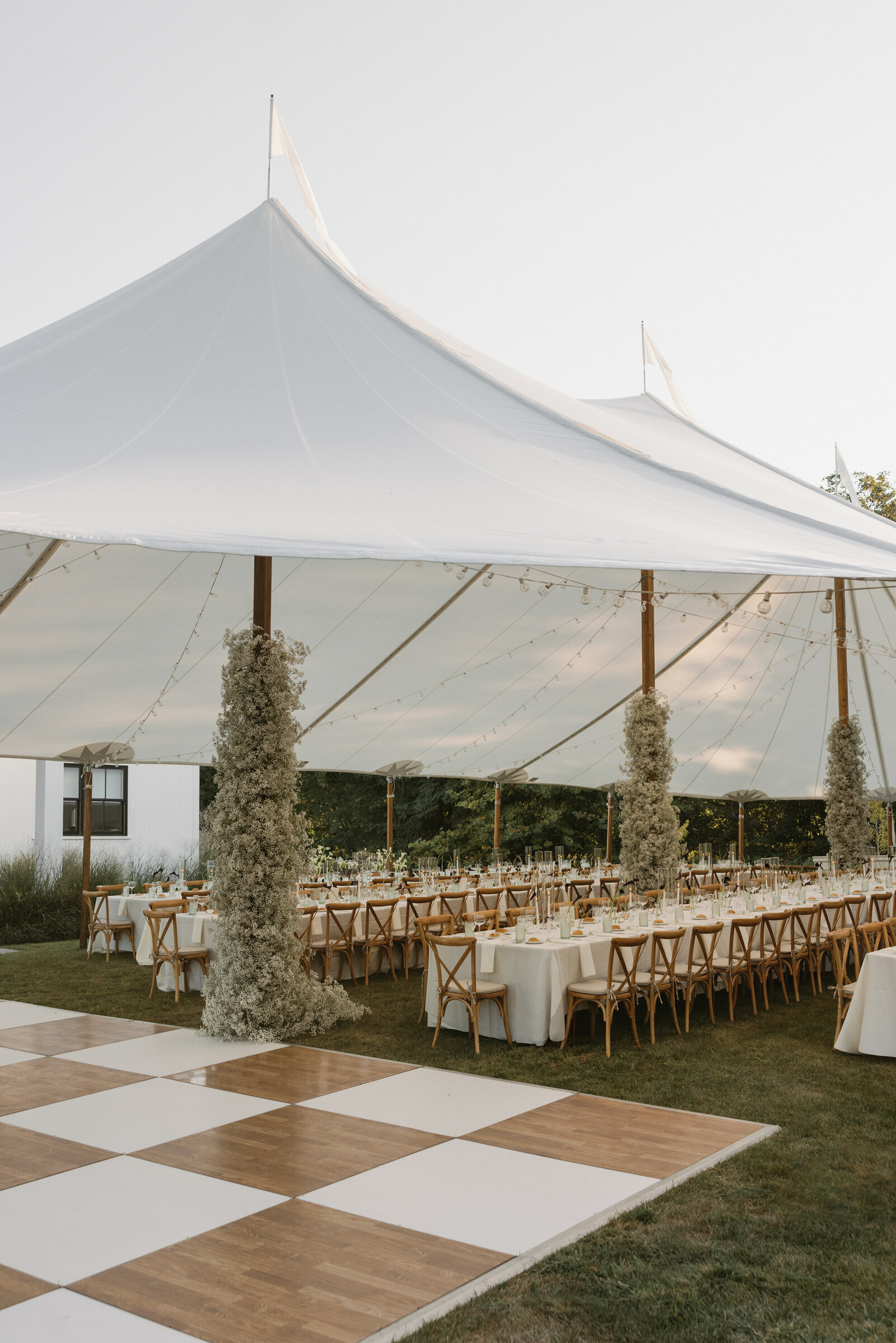 Sailcloth wedding reception tent with baby's breath along tent poles, wooden cross-back chairs lining long white tables, and brown & white checkerboard dance floor 