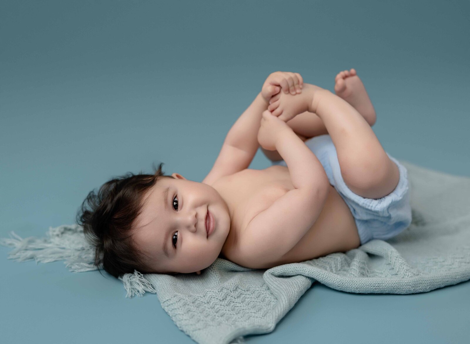 Baby laying on his back with a blue diaper cover on and grabbing his toes, smiling. Photographed at Atlanta baby photo studio.
