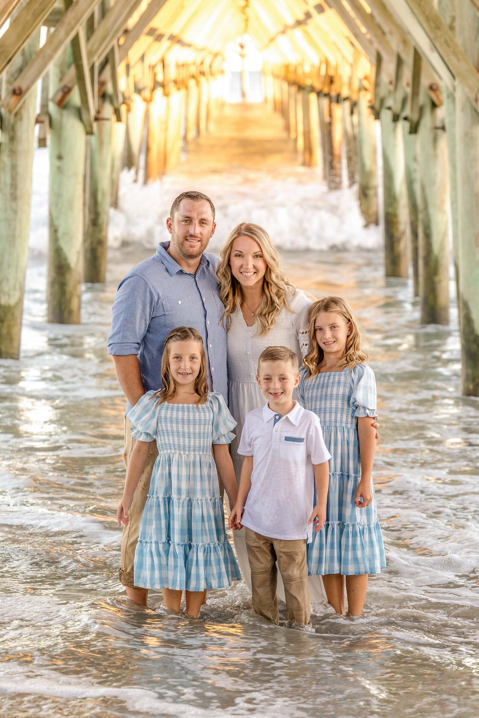 A family of five stands in the water under a pier at sunrise, dressed in coordinating blue and white outfits. The golden light filters through the wooden beams, creating a magical setting. Ideal for North Carolina family photography, capturing enchanting beach moments.