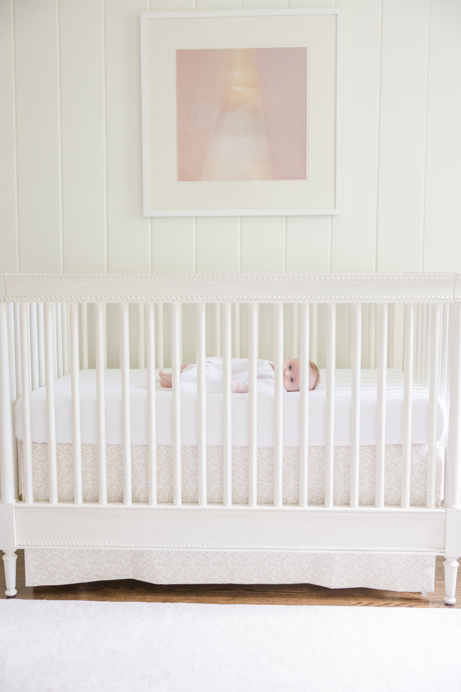 Baby girl laying in a white crib with a pink painting hanging above.