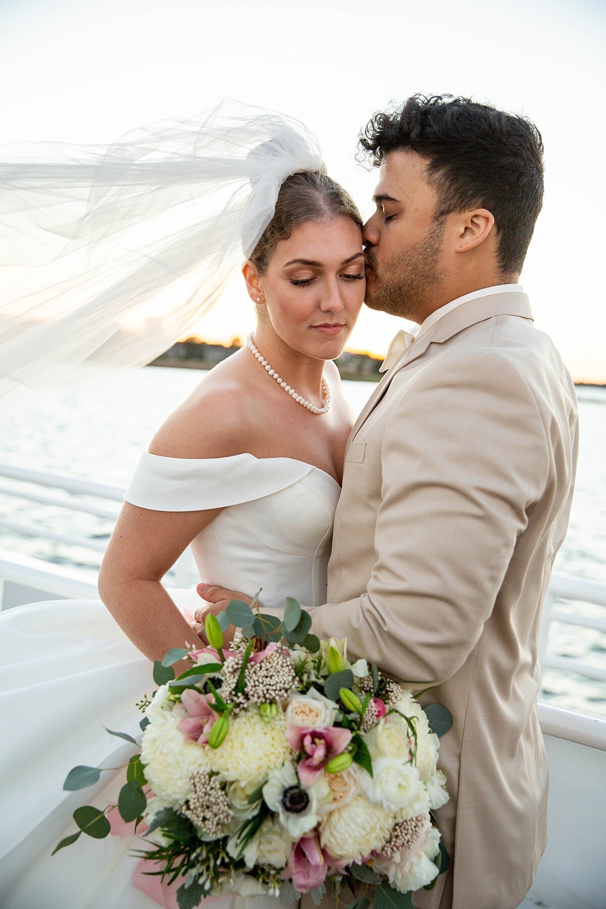 The groom, dressed in a light khaki suit, kisses the bride's temple as she looks down at her bouquet of white, ivory and dark pink flowers with her white dress and veil blowing in the ocean breeze