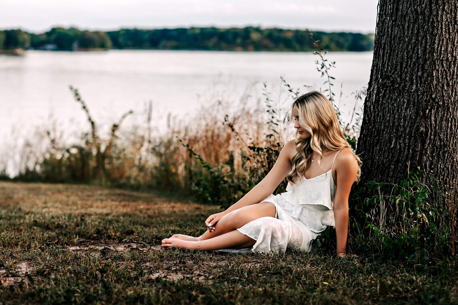 A woman with long blond hair sits barefoot under a tree near a lake, wearing a white dress. The background features calm water and a distant shoreline with trees. The atmosphere is peaceful and serene.