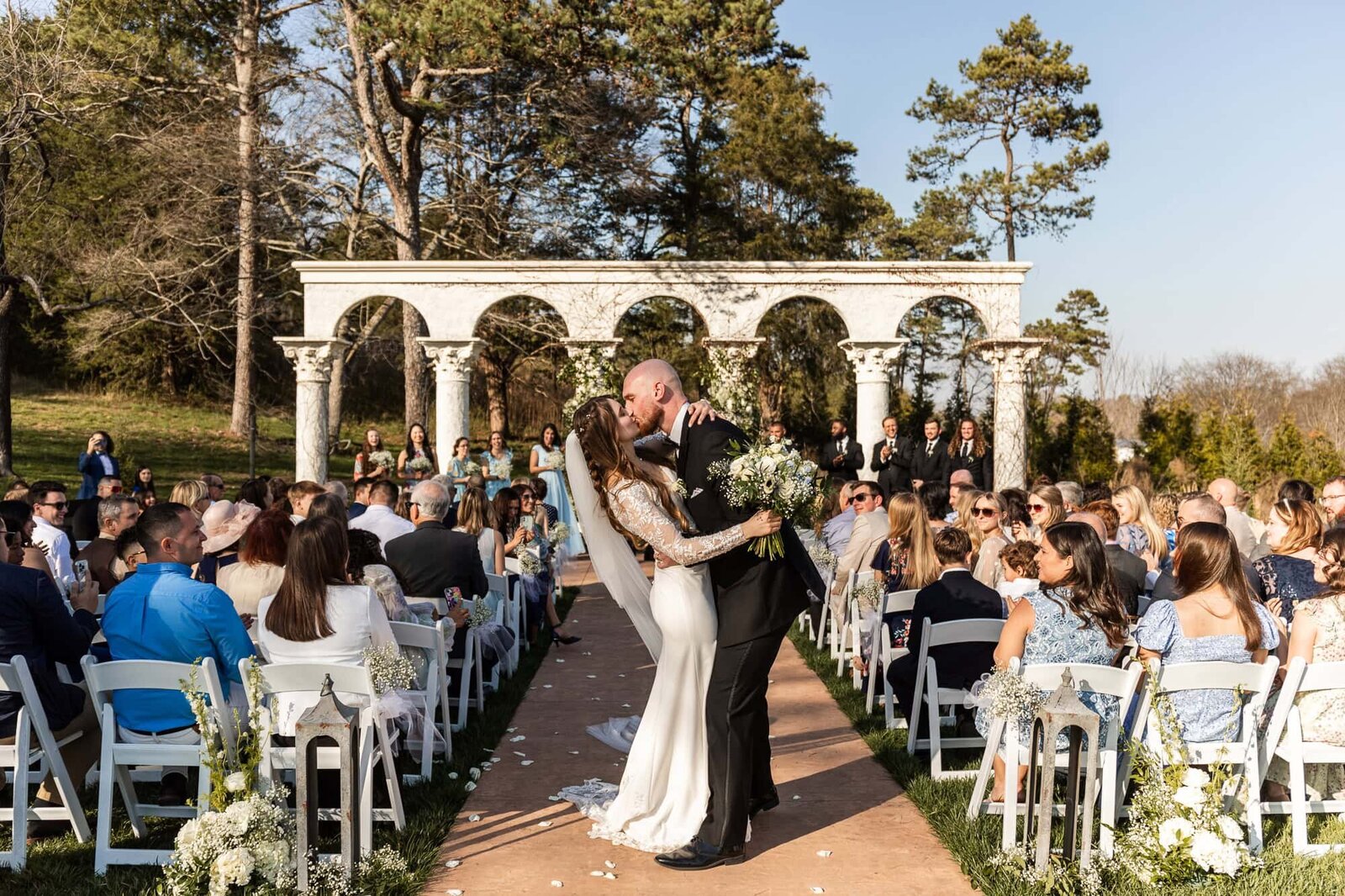 The bride and groom share a celebratory kiss at the end of the aisle, surrounded by cheering guests. The sun casts a golden glow over the elegant Greco-Roman-style columns framing their ceremony backdrop. The bride’s fitted lace wedding dress flows behind her as she clutches a bouquet of white florals, while the groom leans in, dressed in a sharp black tux