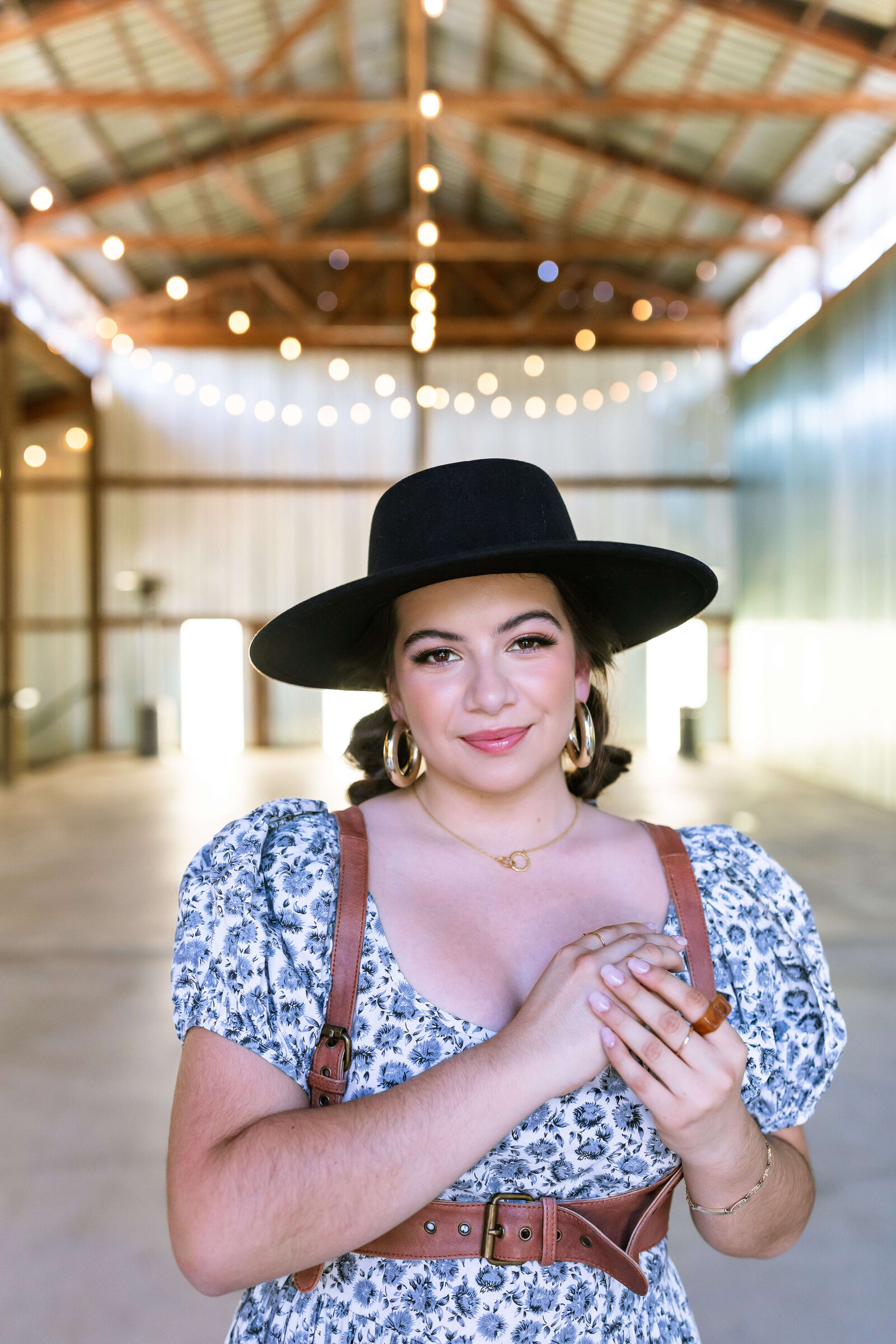 dark haired girl wearing black hat, brown belt, and blue floral dress with bubble braids in a warehouse