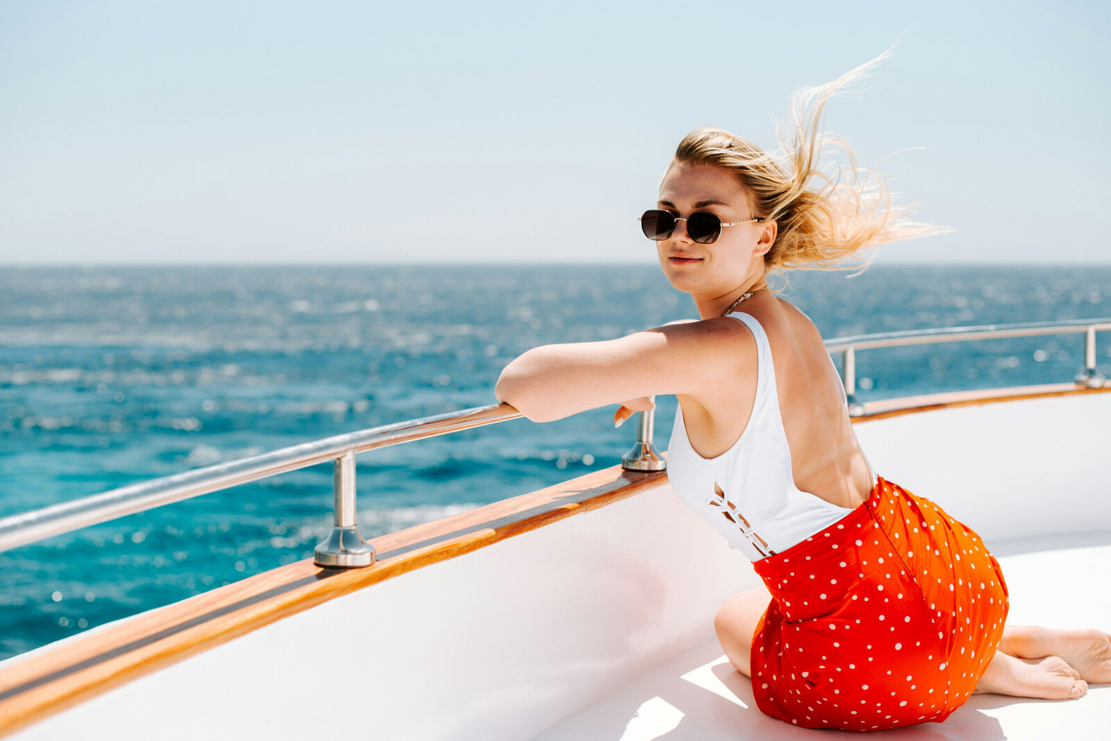 A woman sitting on the deck of a boat leaning on a railing and looking back, with the ocean and clear sky in the background